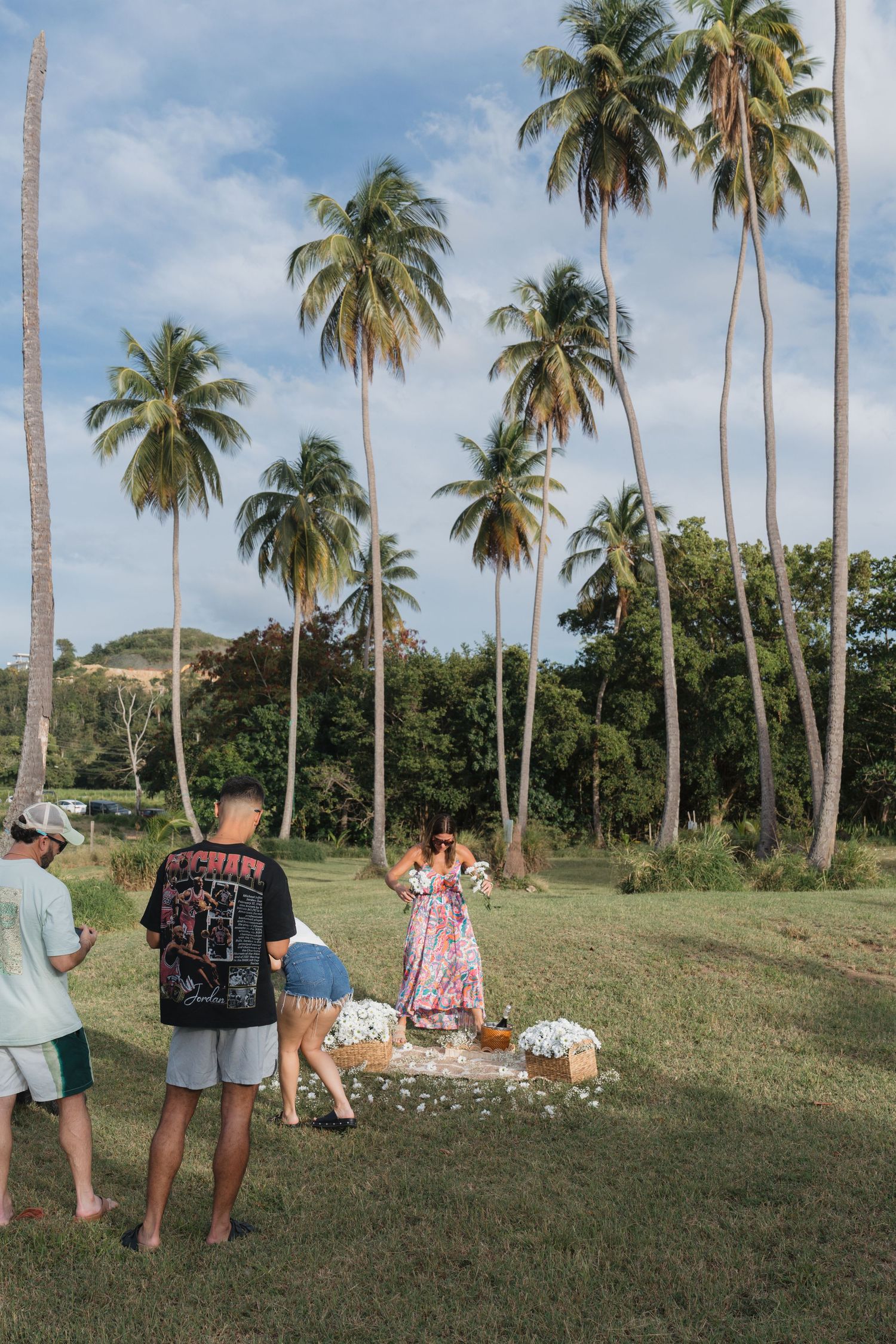 People gathered under tall palm trees on a tropical lawn, with flowers arranged on the ground during an outdoor celebration.