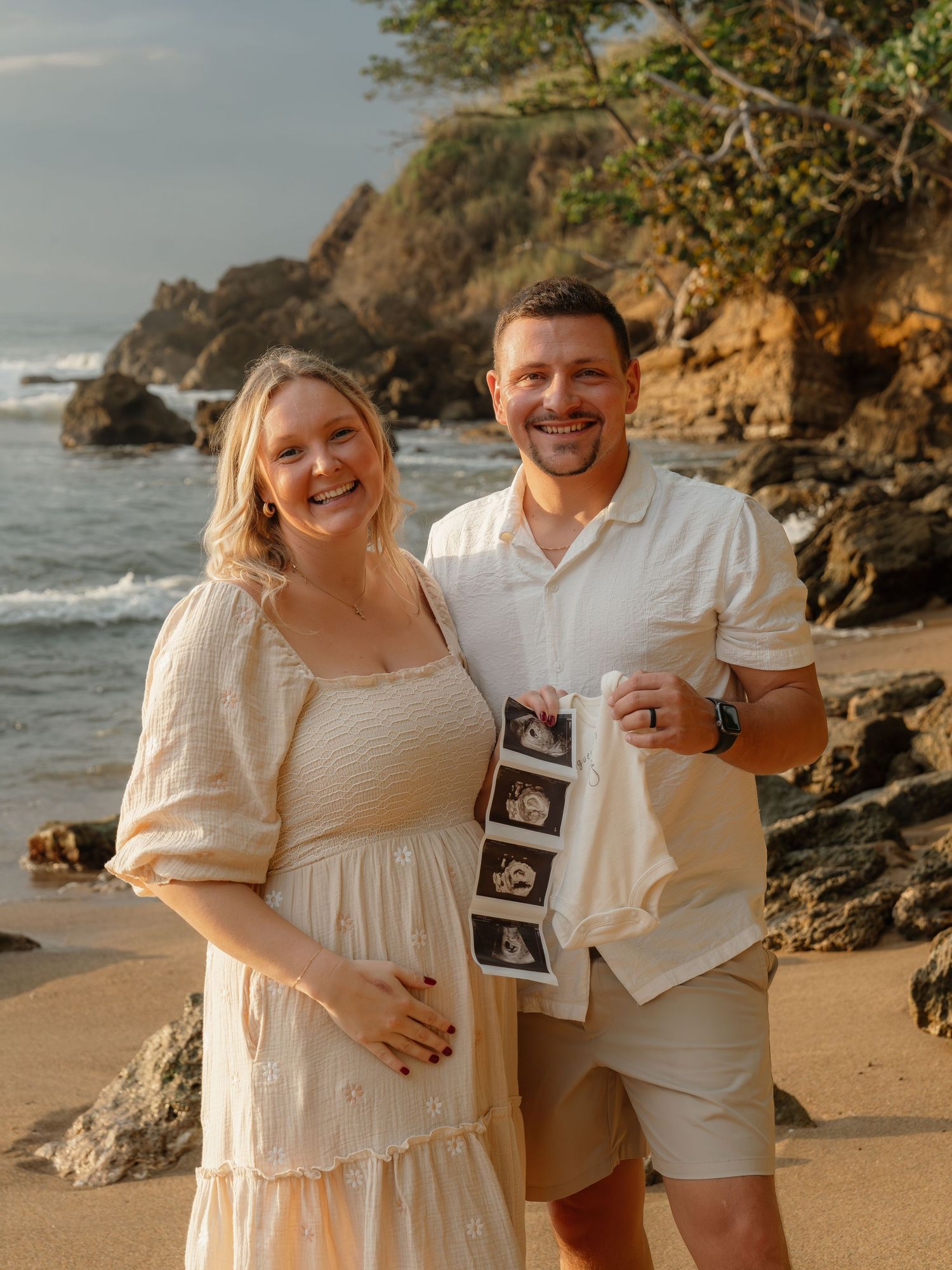 Pregnant couple smiling on a beach holding ultrasound photos, with rocky cliffs and ocean waves in the background.