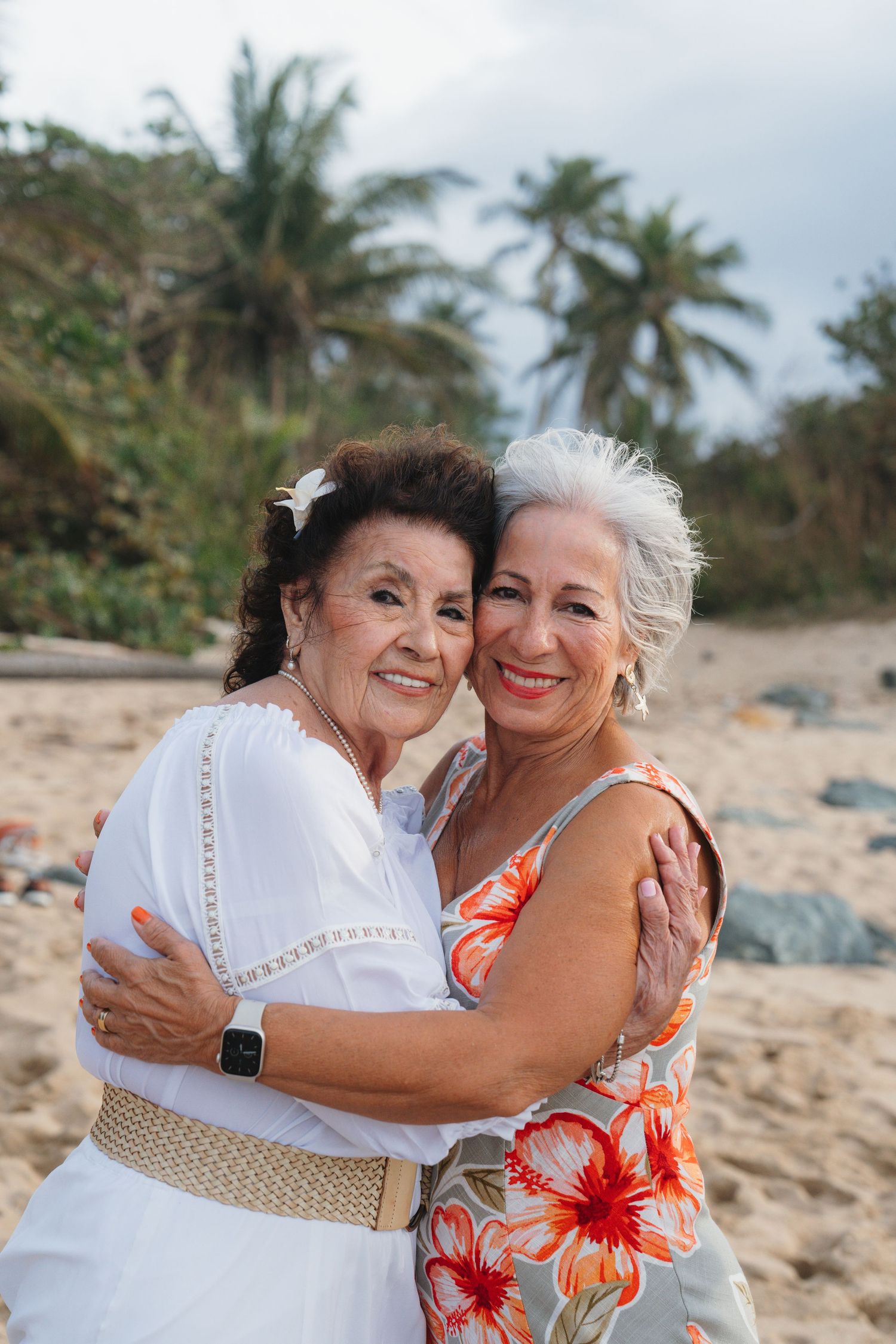 Two senior women hugging and smiling on a tropical beach, one in white dress, other in floral sundress.