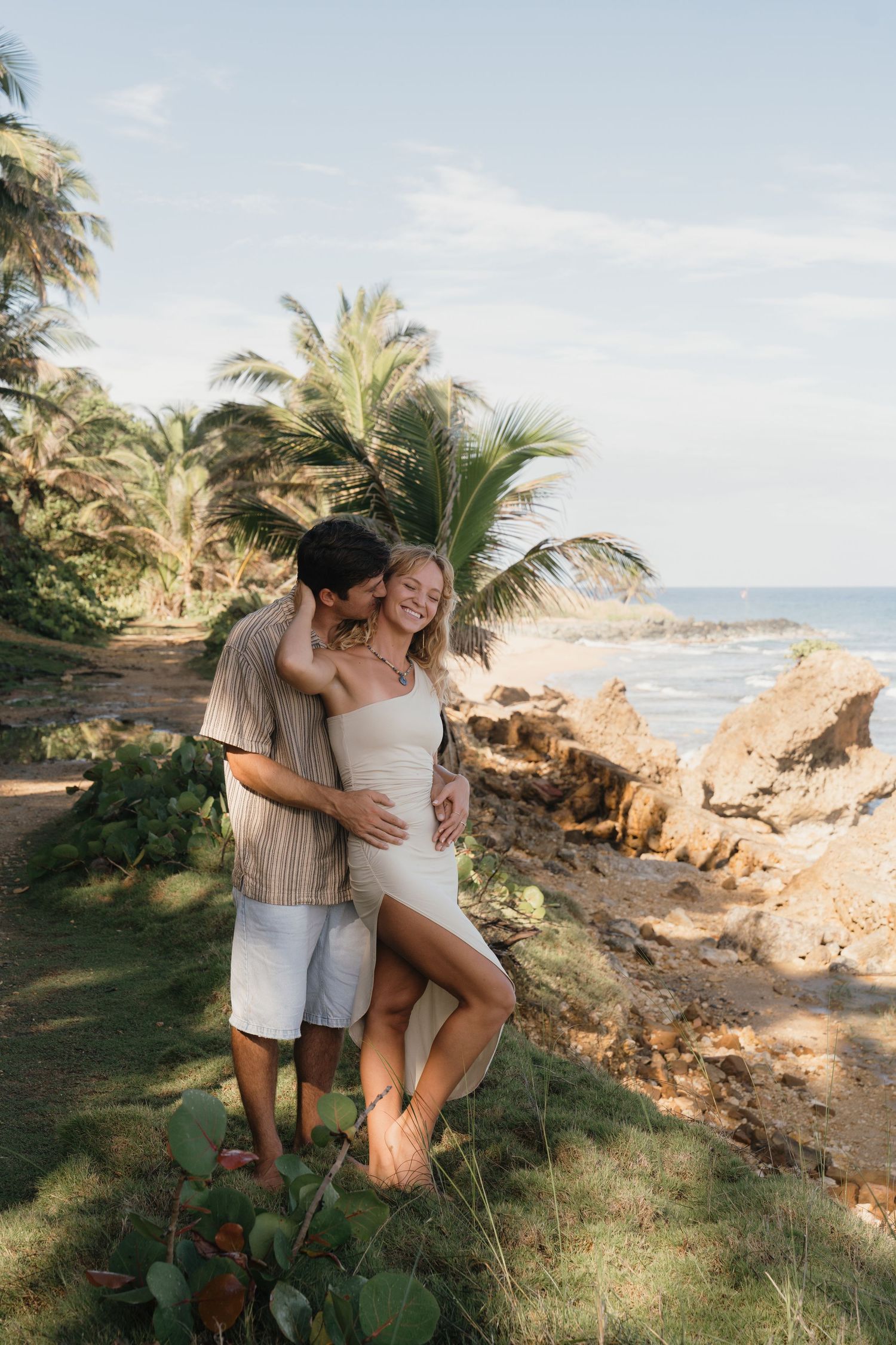 A couple embraces on a tropical beach path surrounded by palm trees and rocky shoreline on a sunny day.