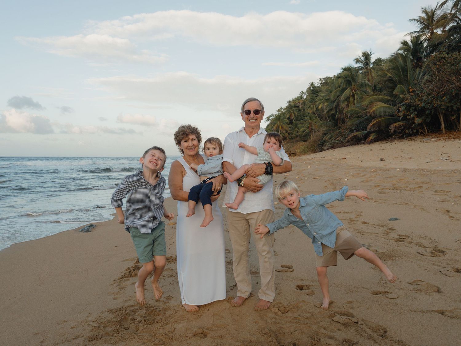 Family of six posing happily on a tropical beach with palm trees and ocean waves in the background.