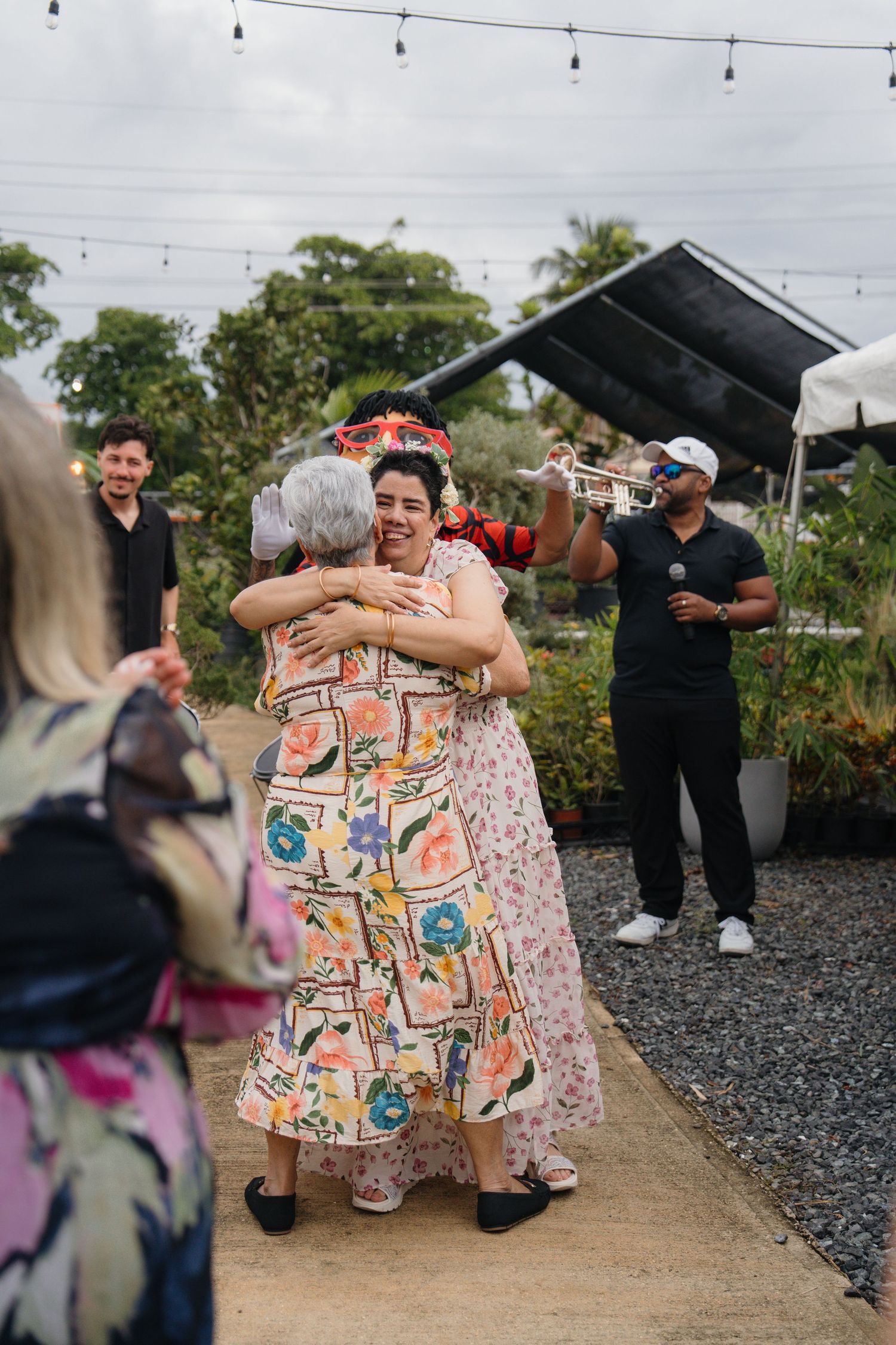 Two women in floral dresses share a warm hug at an outdoor garden party with string lights and live music.