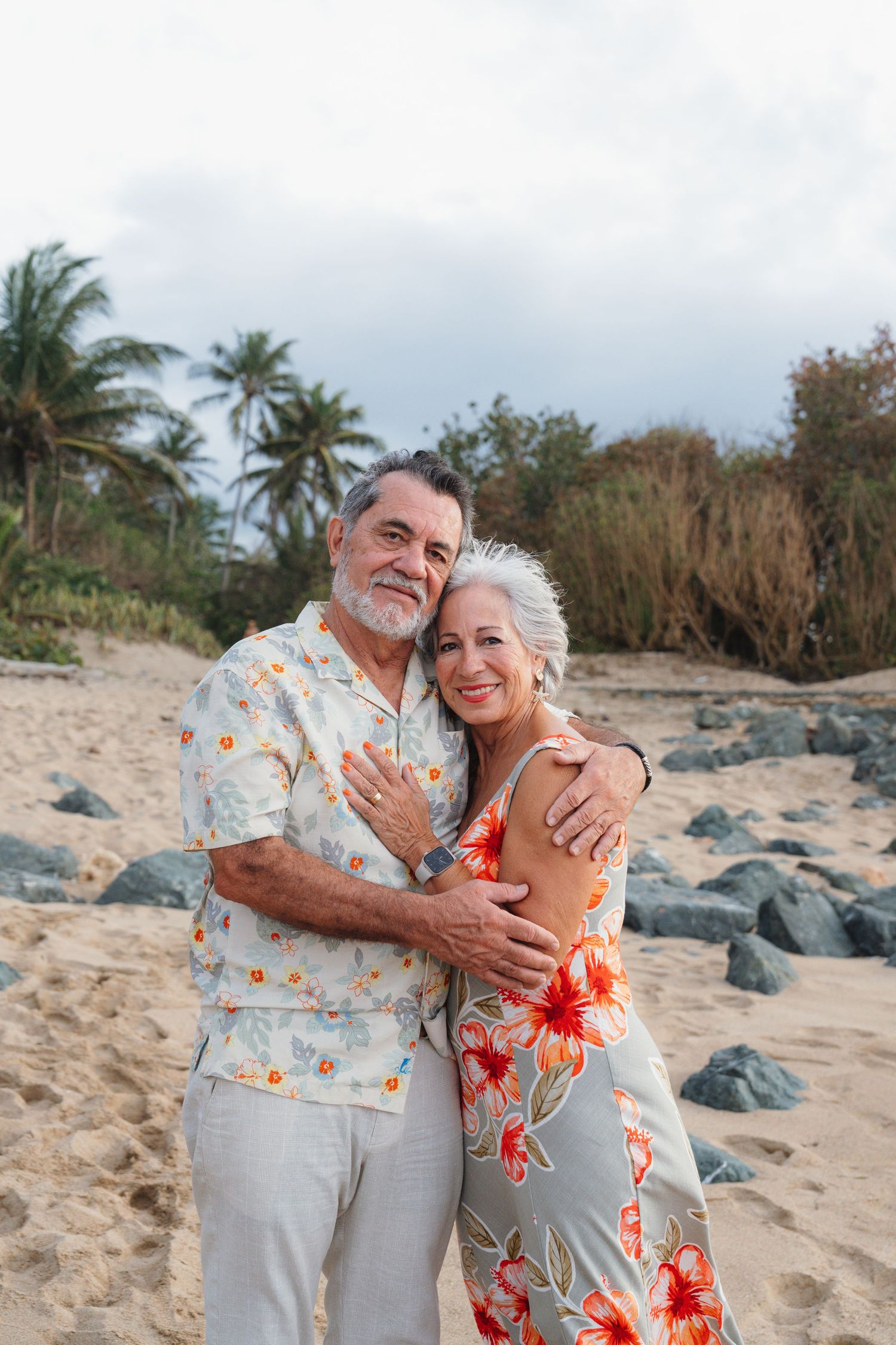 A happy senior couple embraces on a tropical beach, wearing matching floral outfits with palm trees in the background.