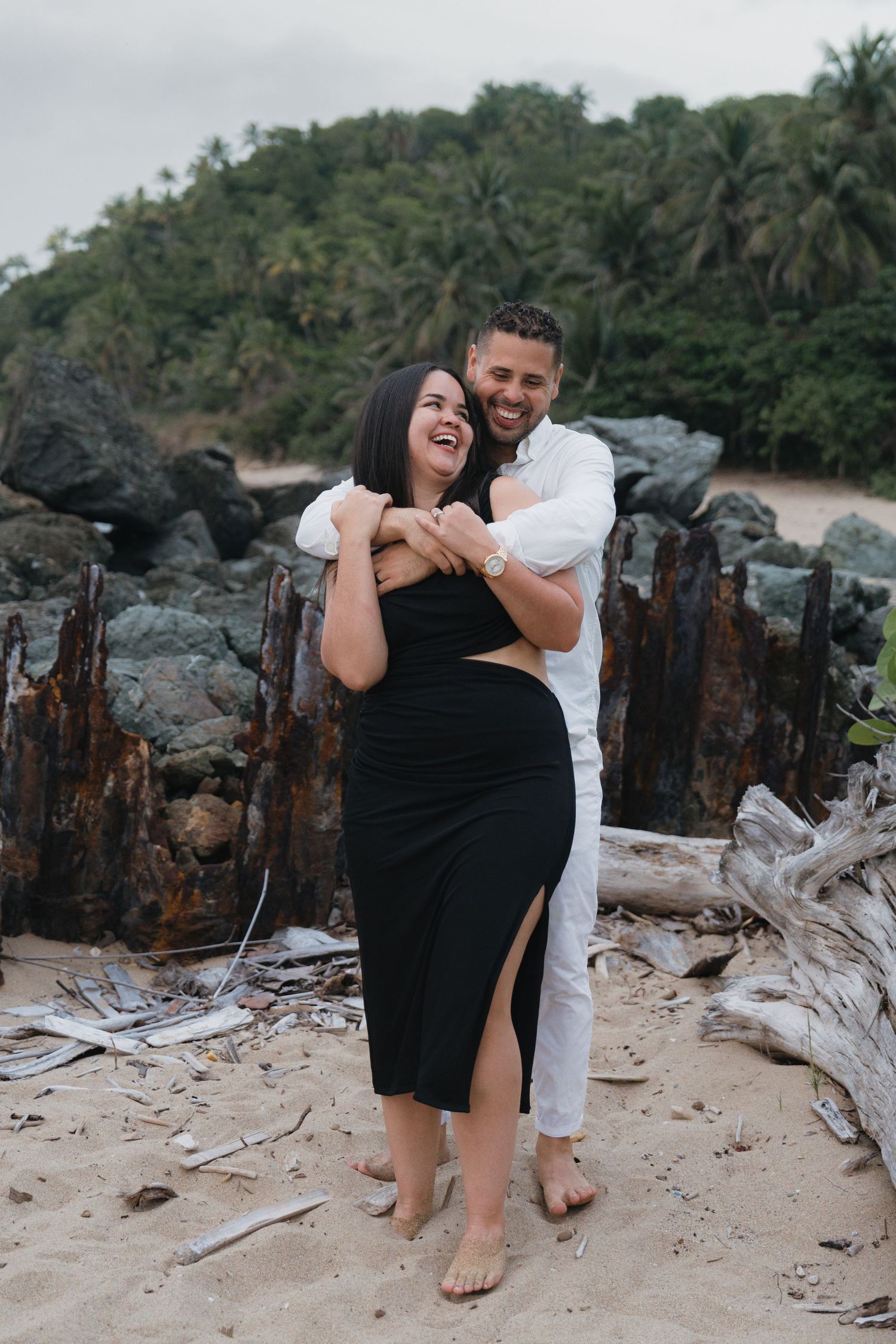 Happy couple embracing on a tropical beach, woman in black dress, man in white outfit, with rocks and palm trees behind.