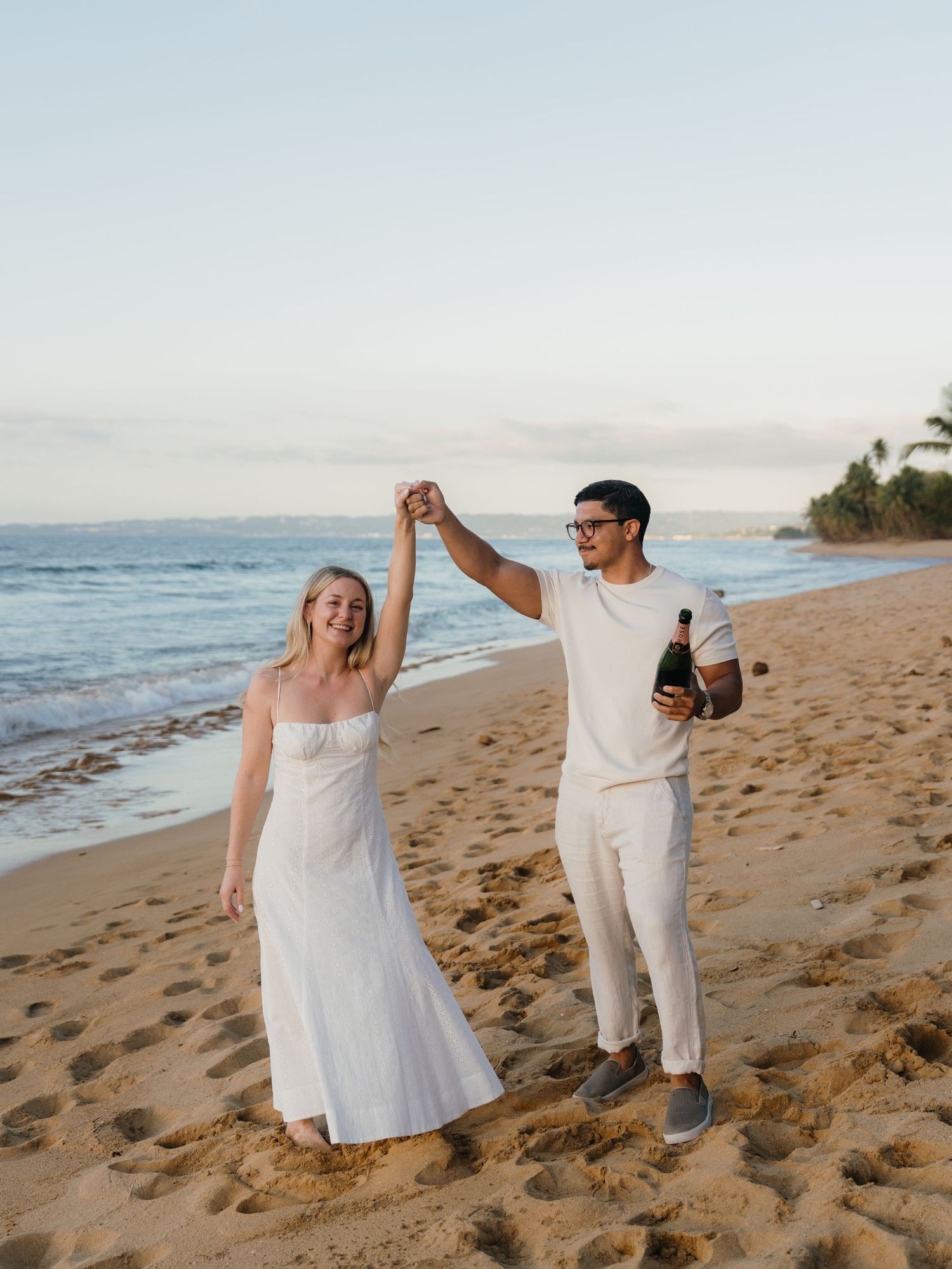 A joyful couple in white outfits dancing on a sandy beach at sunset, holding hands with a champagne bottle.