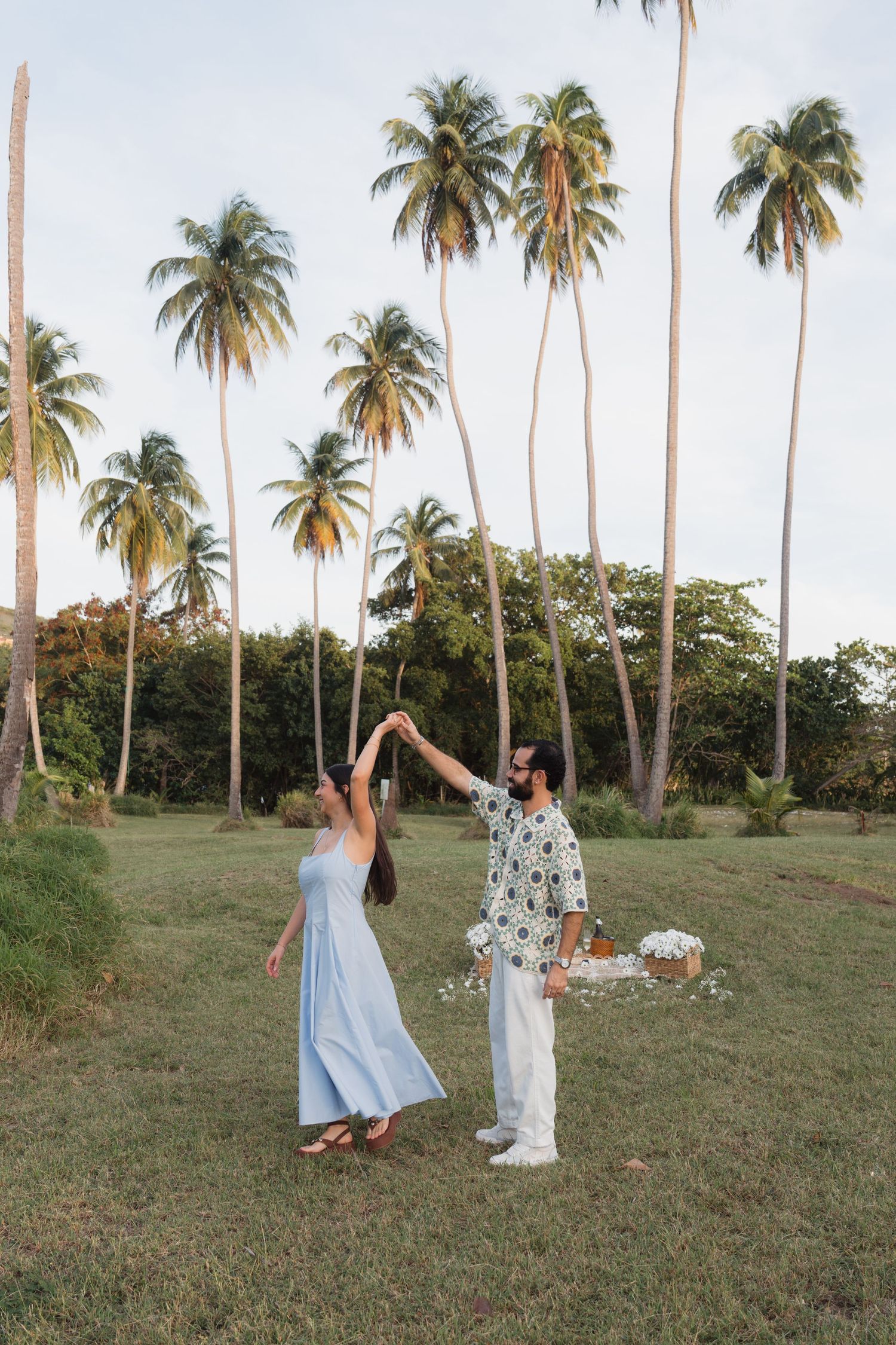 Couple dancing together on a tropical lawn surrounded by tall palm trees, woman in light blue dress, man in floral shirt.