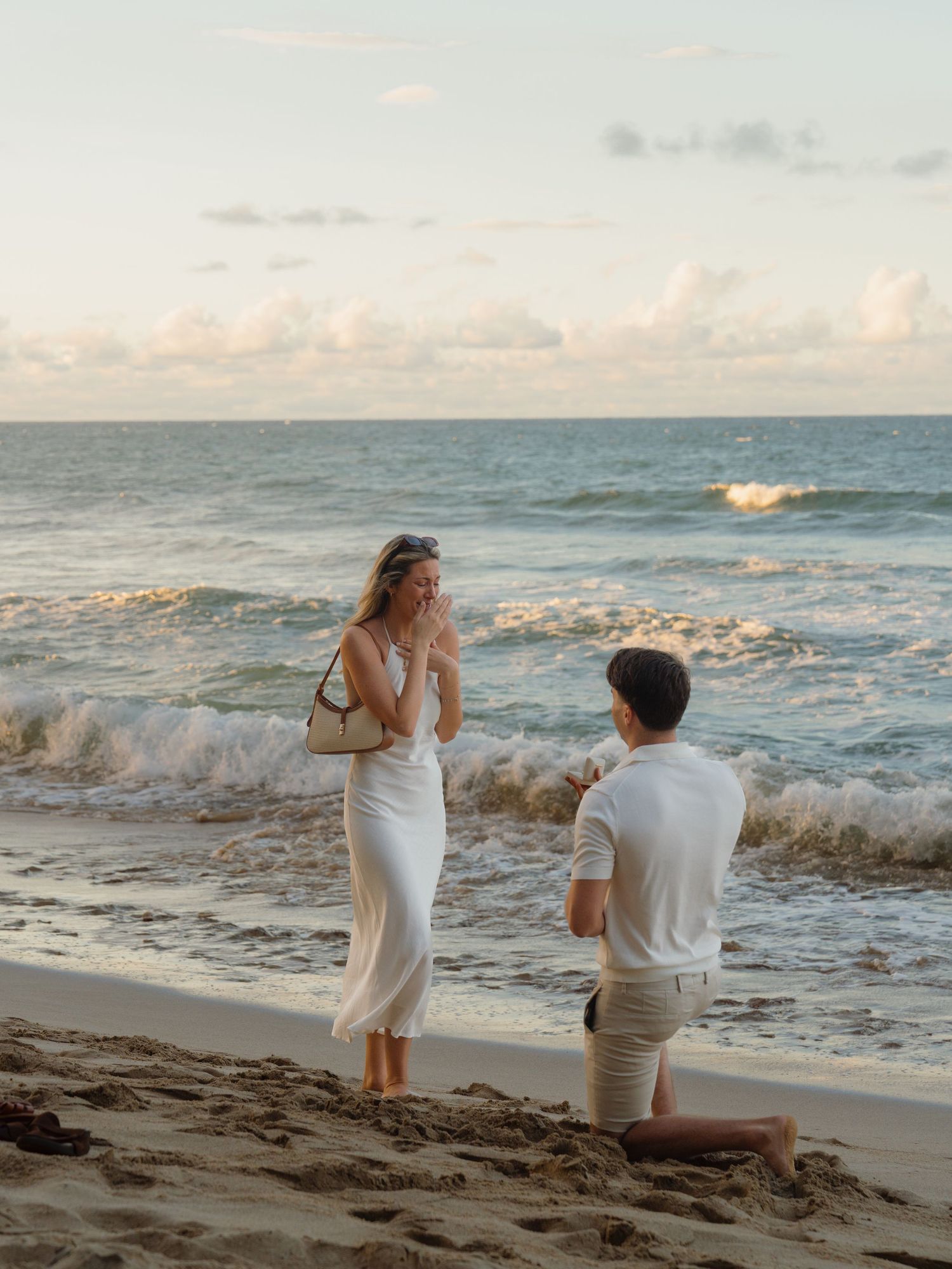 Man kneeling on sandy beach proposing to woman in white dress at sunset by ocean waves.