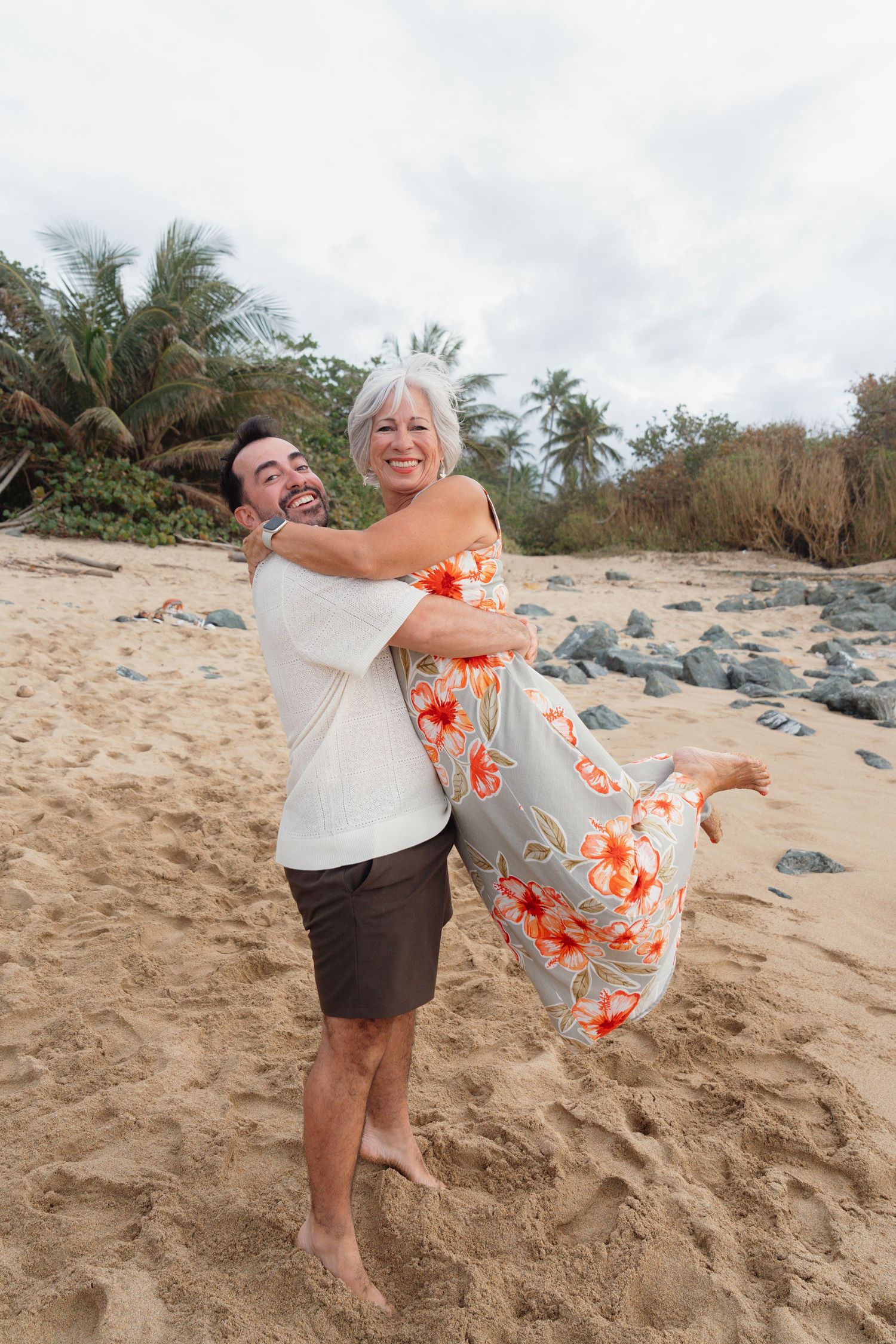 Happy couple embracing on a tropical beach, man lifting woman in floral dress, palm trees and sand in background.