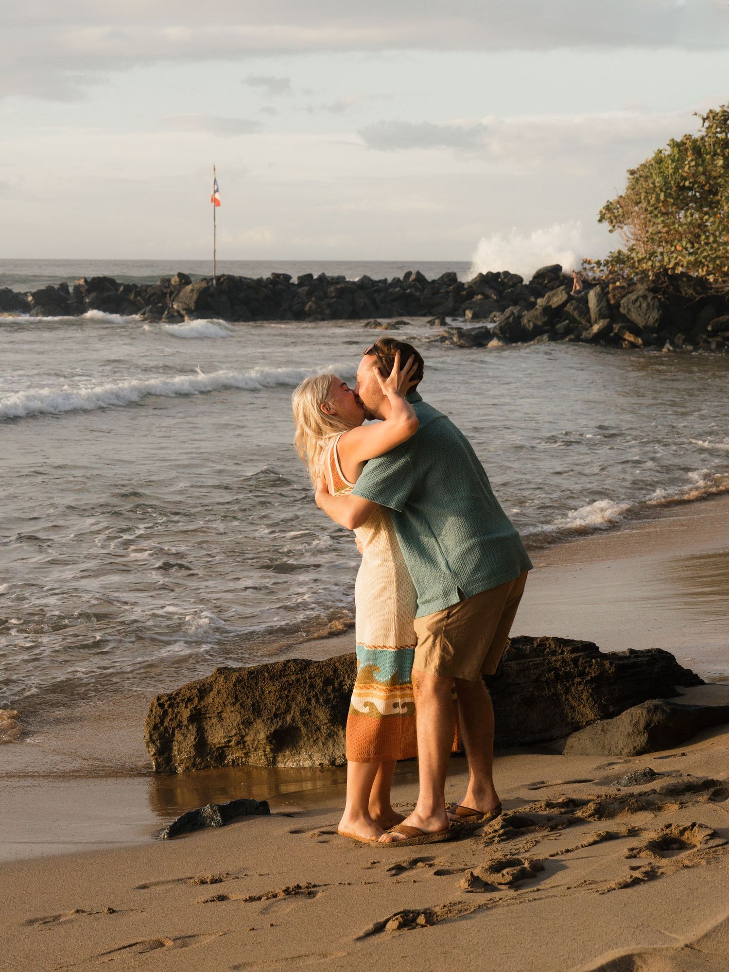 Couple sharing a romantic kiss on a sandy beach at sunset with ocean waves and rocks in the background.