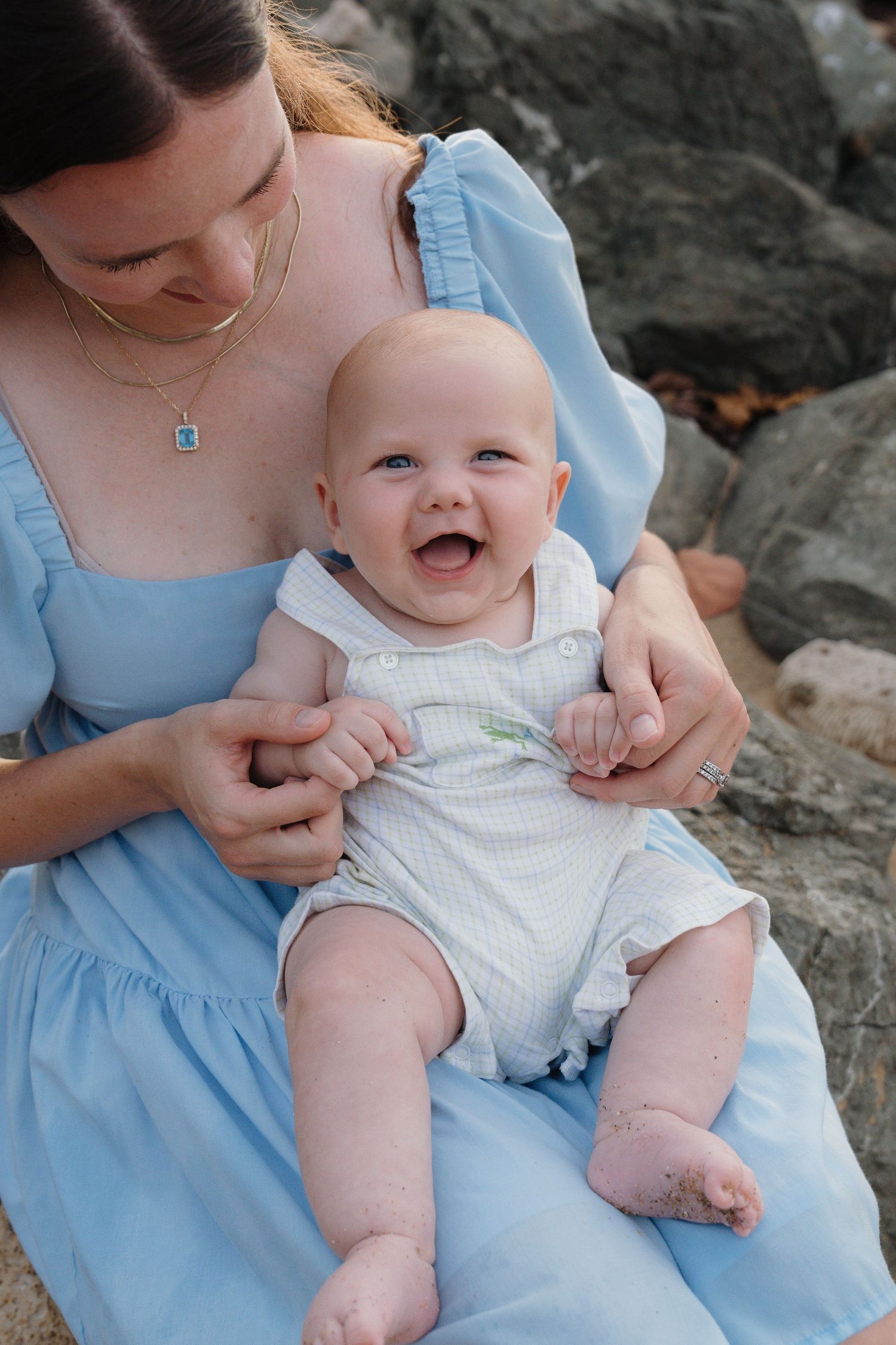 A smiling baby in white romper sits on mother's lap in blue dress near rocks at the beach.