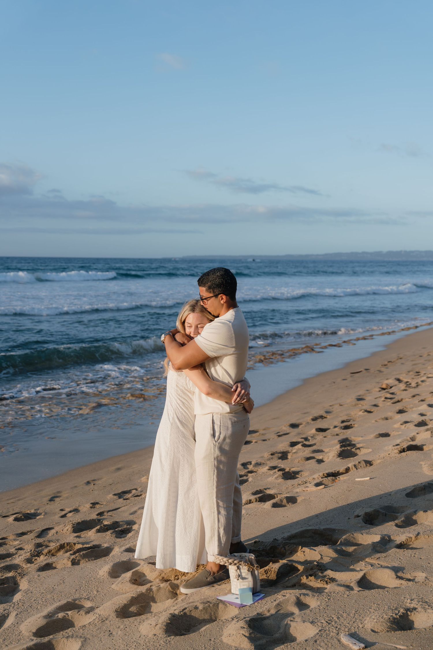 Couple embracing on a sandy beach at sunset, wearing white outfits, with ocean waves in the background.