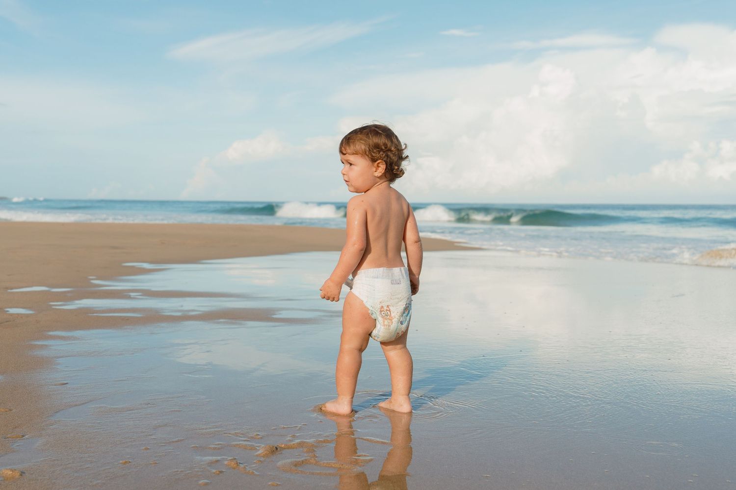 Toddler in white diaper walking barefoot on sandy beach with ocean waves in background.