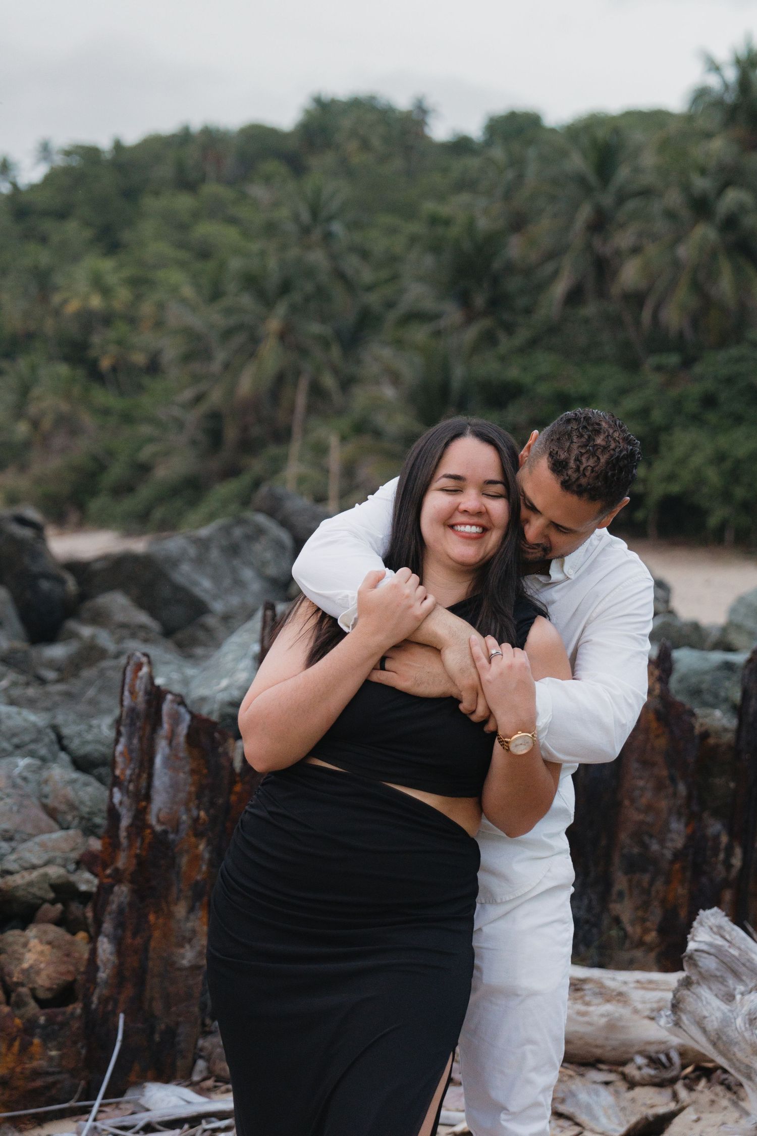 A couple embraces on a rocky beach with lush tropical greenery, the woman in black smiling joyfully.