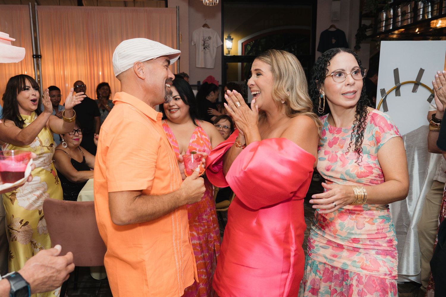 Guests in colorful outfits celebrate at a festive party, with women in pink dresses clapping joyfully.