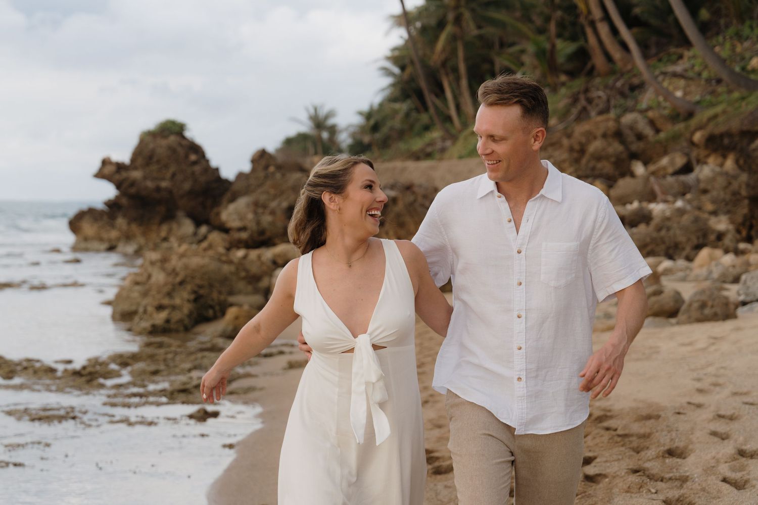 Happy couple walking hand in hand on a tropical beach, woman in white dress, man in linen shirt, rocky coastline backdrop.