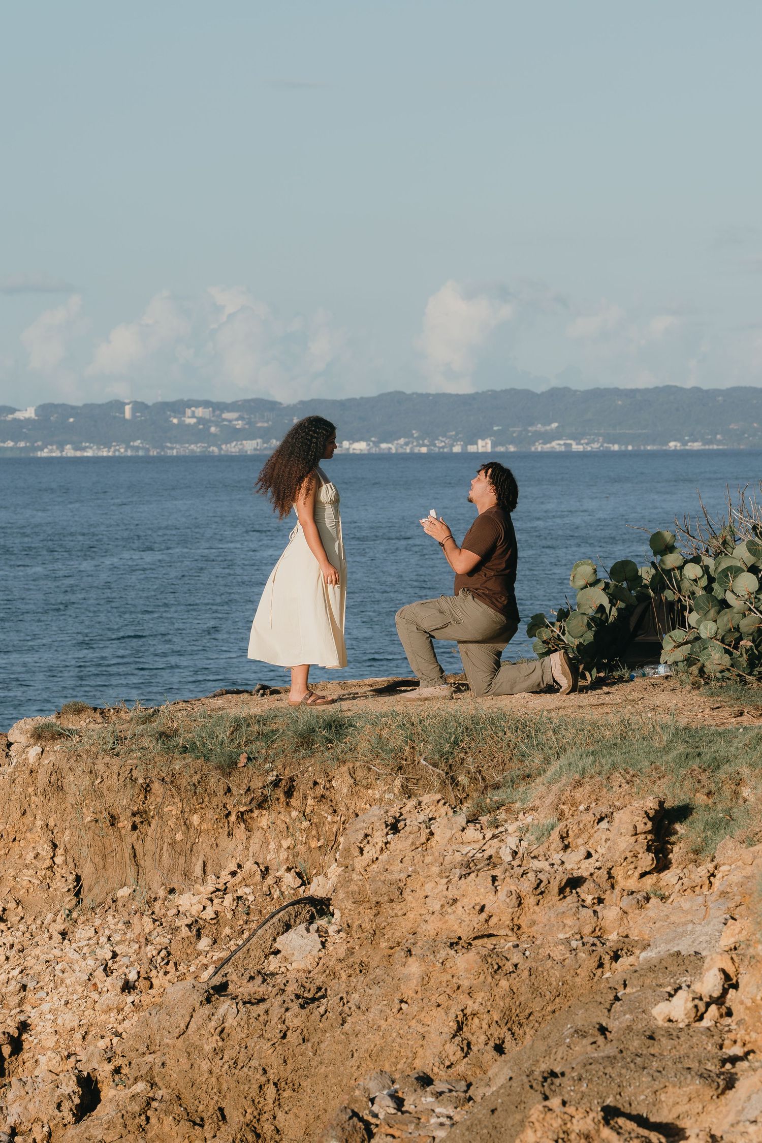 Man proposes on one knee to woman in white dress on rocky coastal cliff with ocean backdrop.