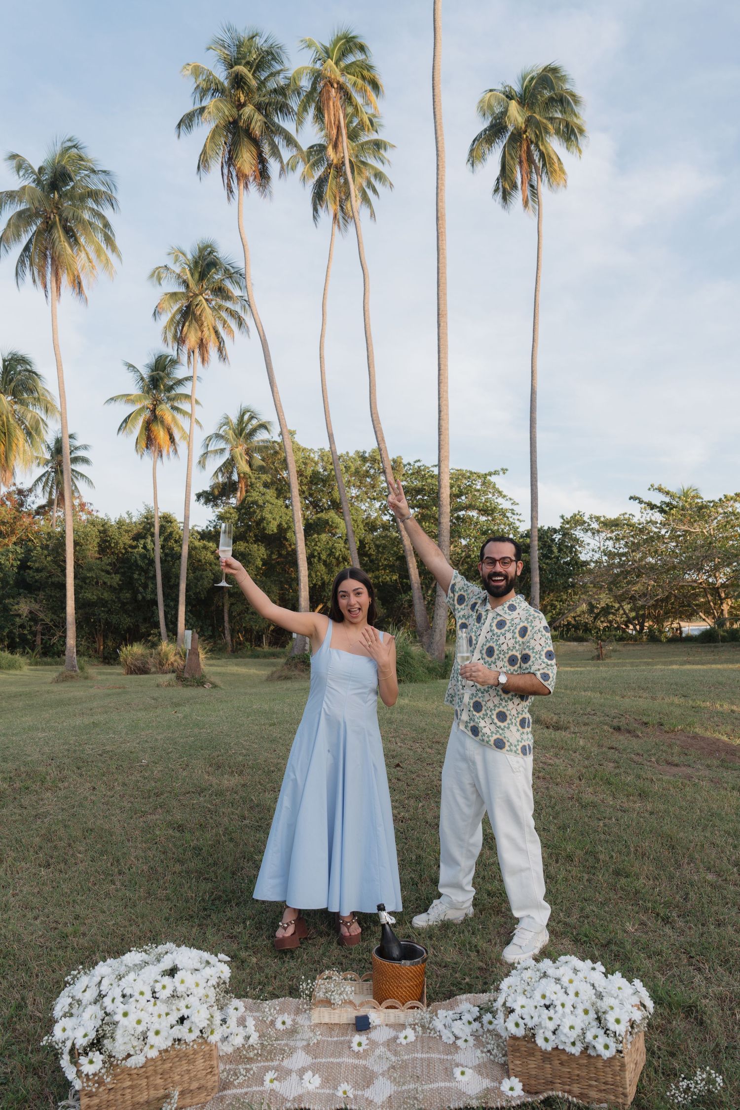 Couple celebrating outdoors under tall palm trees with white flowers and a picnic setup on green grass.