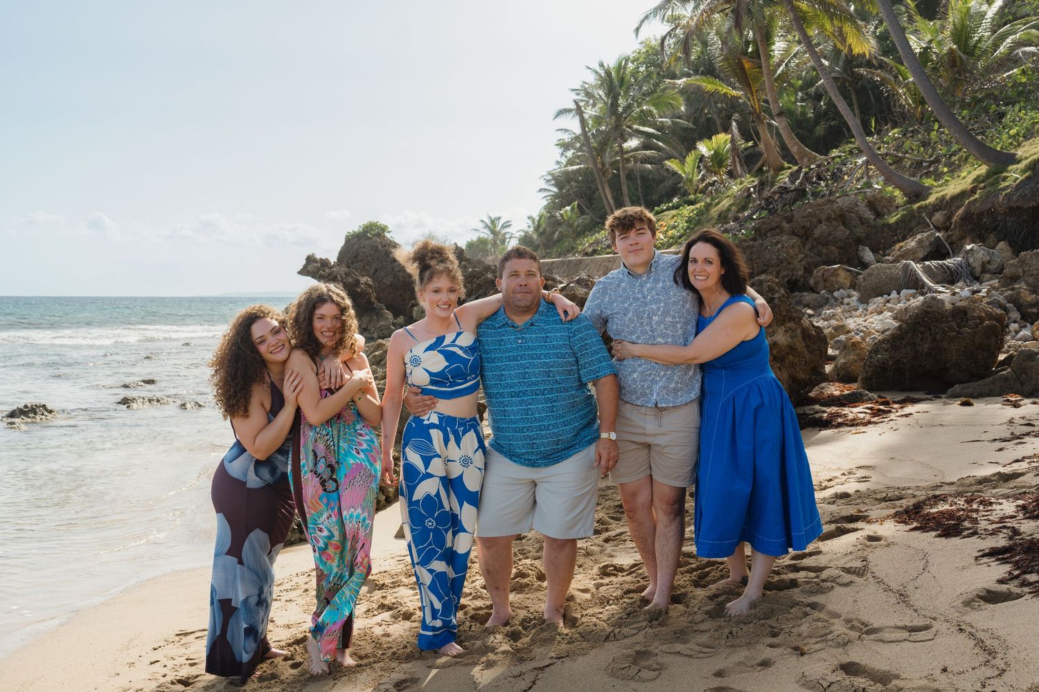 Family of six posing together on a tropical beach with palm trees and rocky shore in the background.
