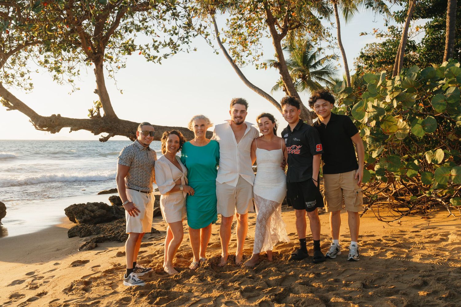 Group of seven people smiling together on a tropical beach with palm trees and ocean waves in the background.