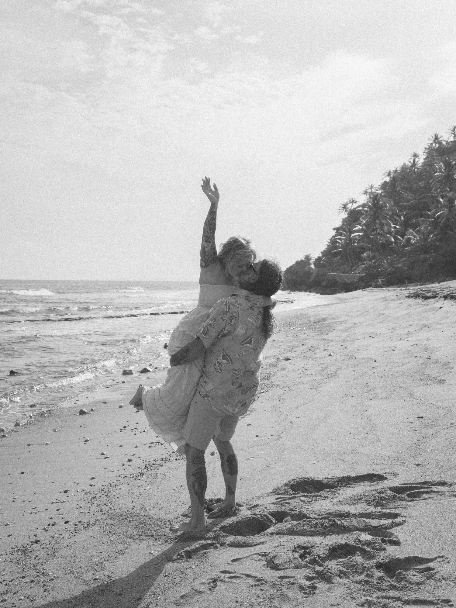 Black and white photo of a couple embracing and dancing joyfully on a sandy beach with ocean waves.