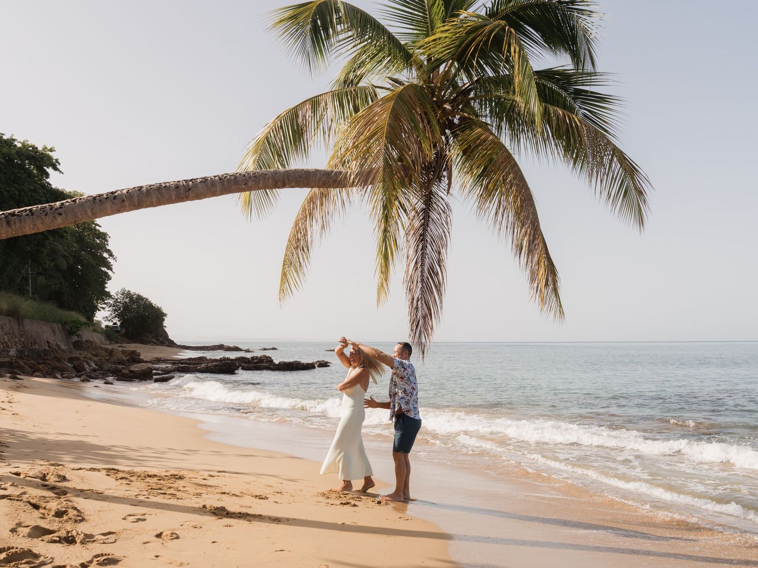 Couple dancing on a tropical beach under a leaning palm tree with calm ocean waves in the background.