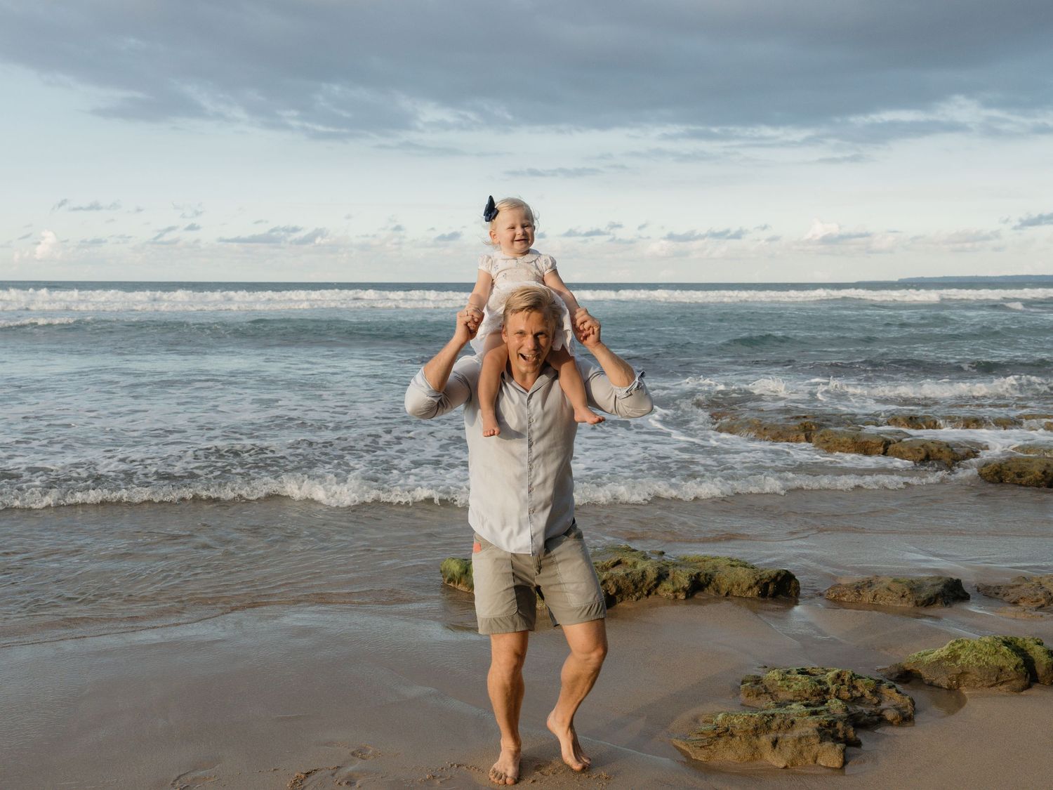 A father carries his toddler on his shoulders while walking barefoot on a sandy beach with ocean waves behind them.