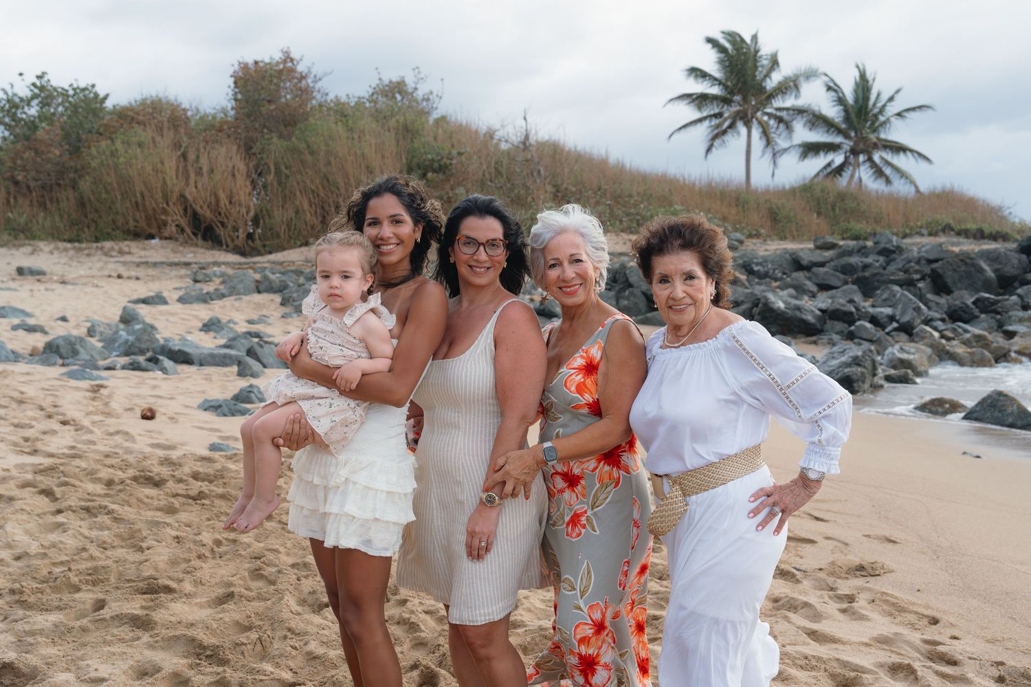 Five women pose together on a sandy beach with palm trees and rocks, wearing white and floral outfits.