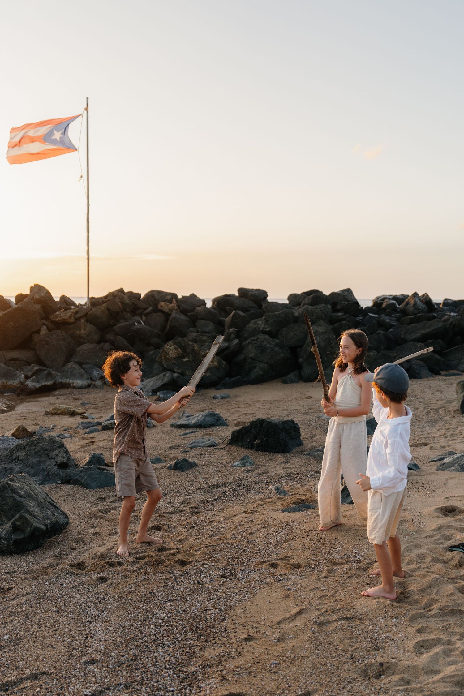 Three children play with sticks on a sandy beach at sunset, with a Puerto Rico flag waving in the background.