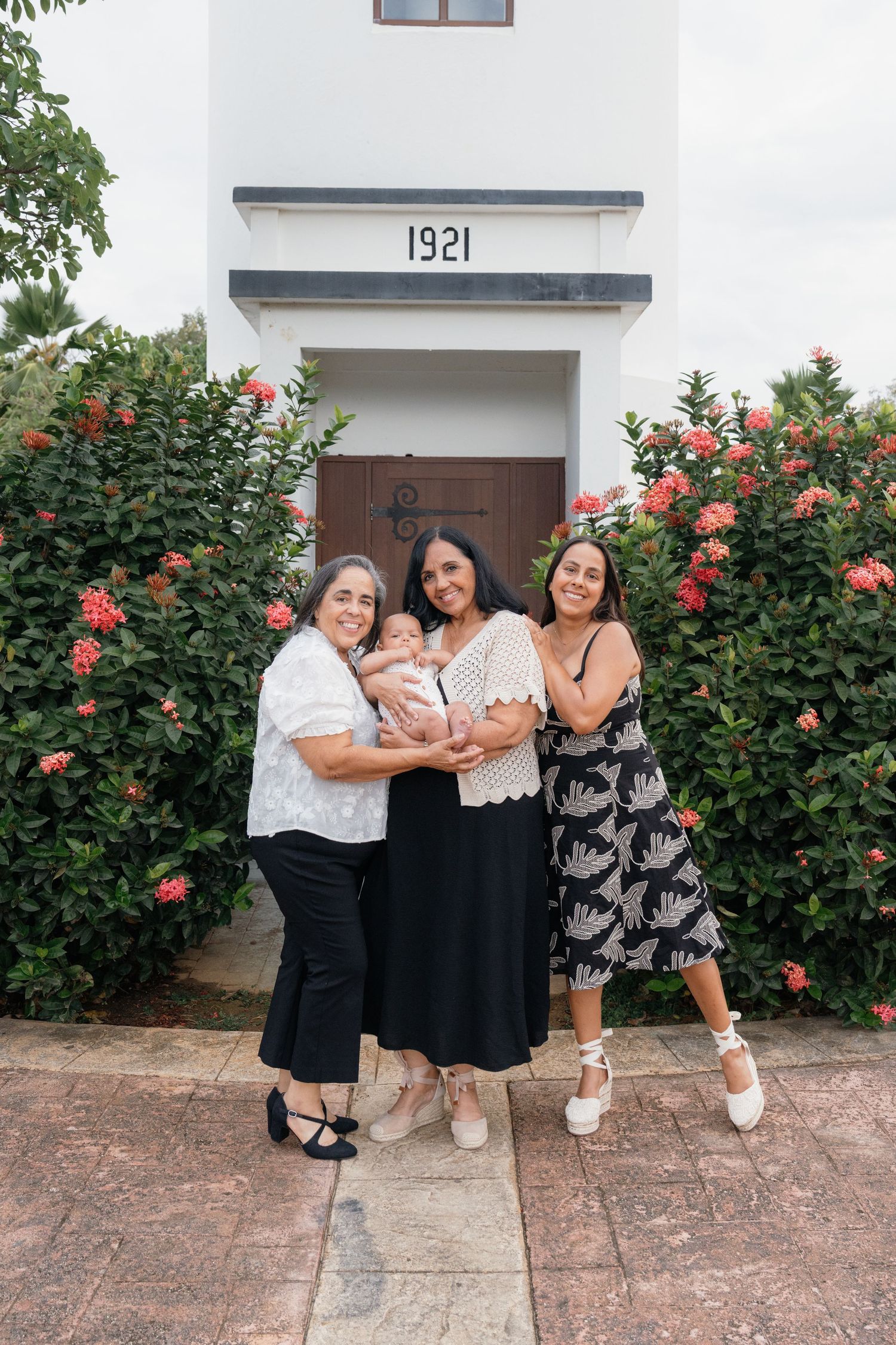 Three women and a baby pose smiling in front of a white building numbered 1921 with lush flowering bushes.