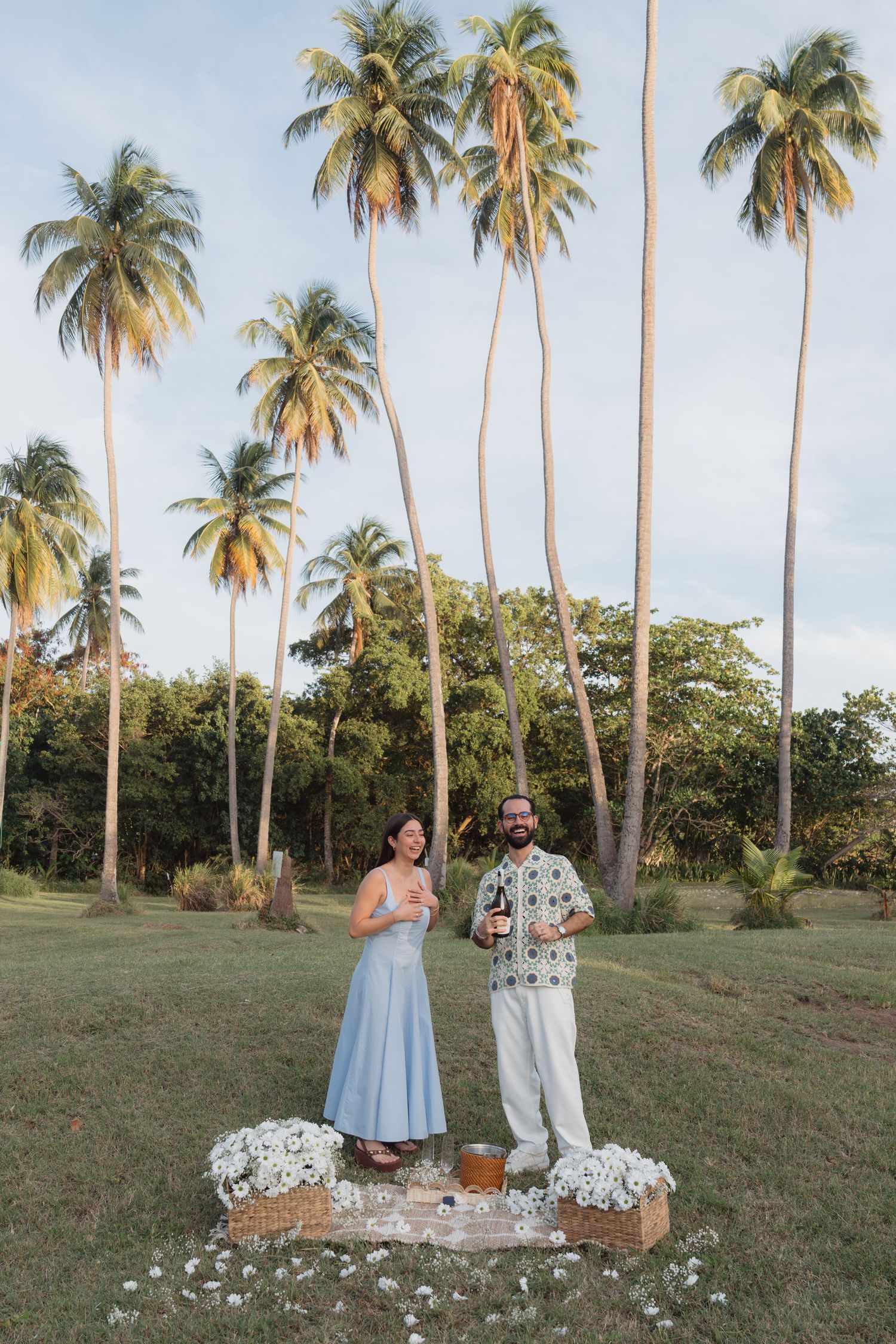 Couple celebrating outdoors surrounded by tall palm trees, standing on a decorated platform with white flowers in a tropical setting.