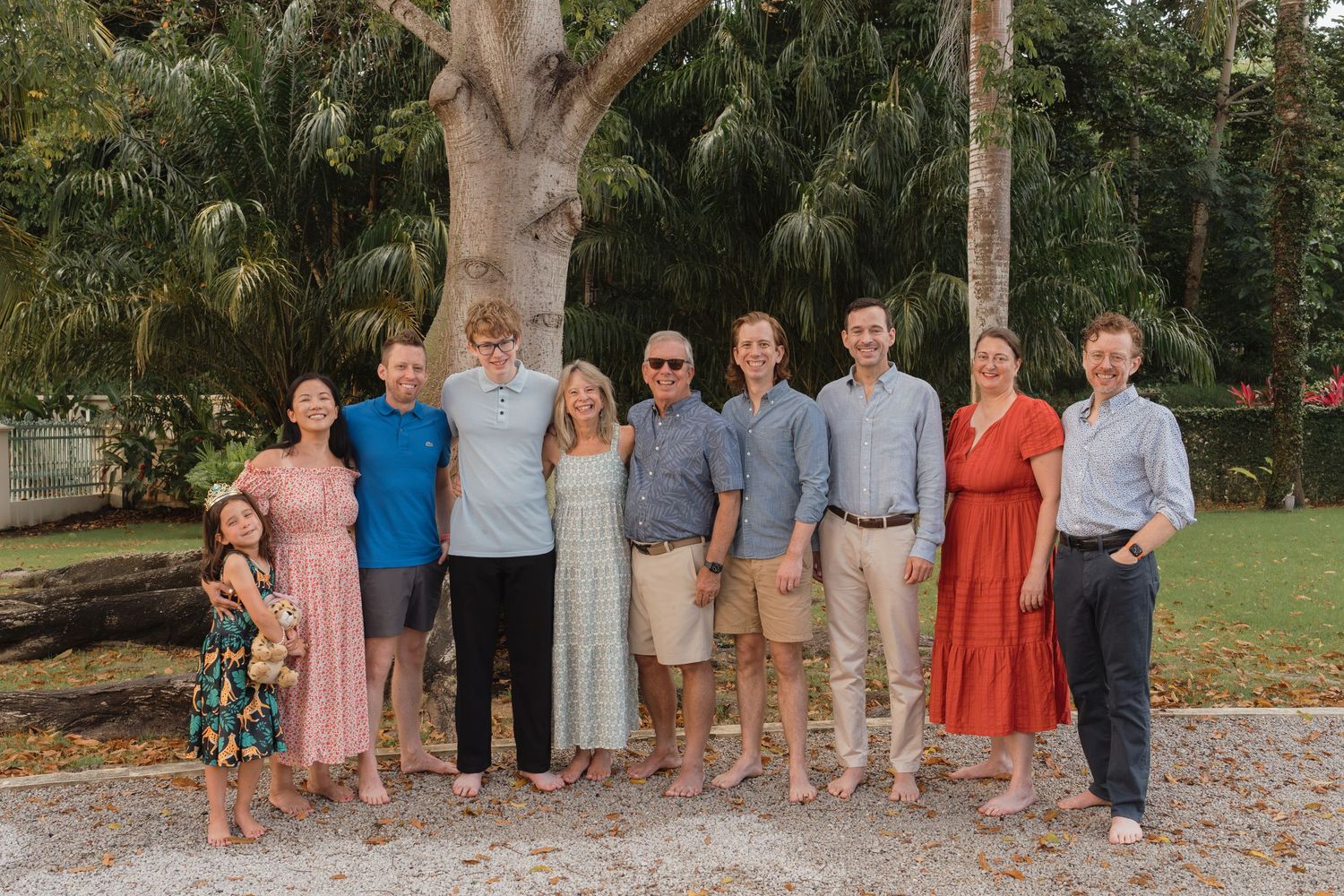 Large family group portrait of ten people smiling outdoors near a large tree in a tropical setting.