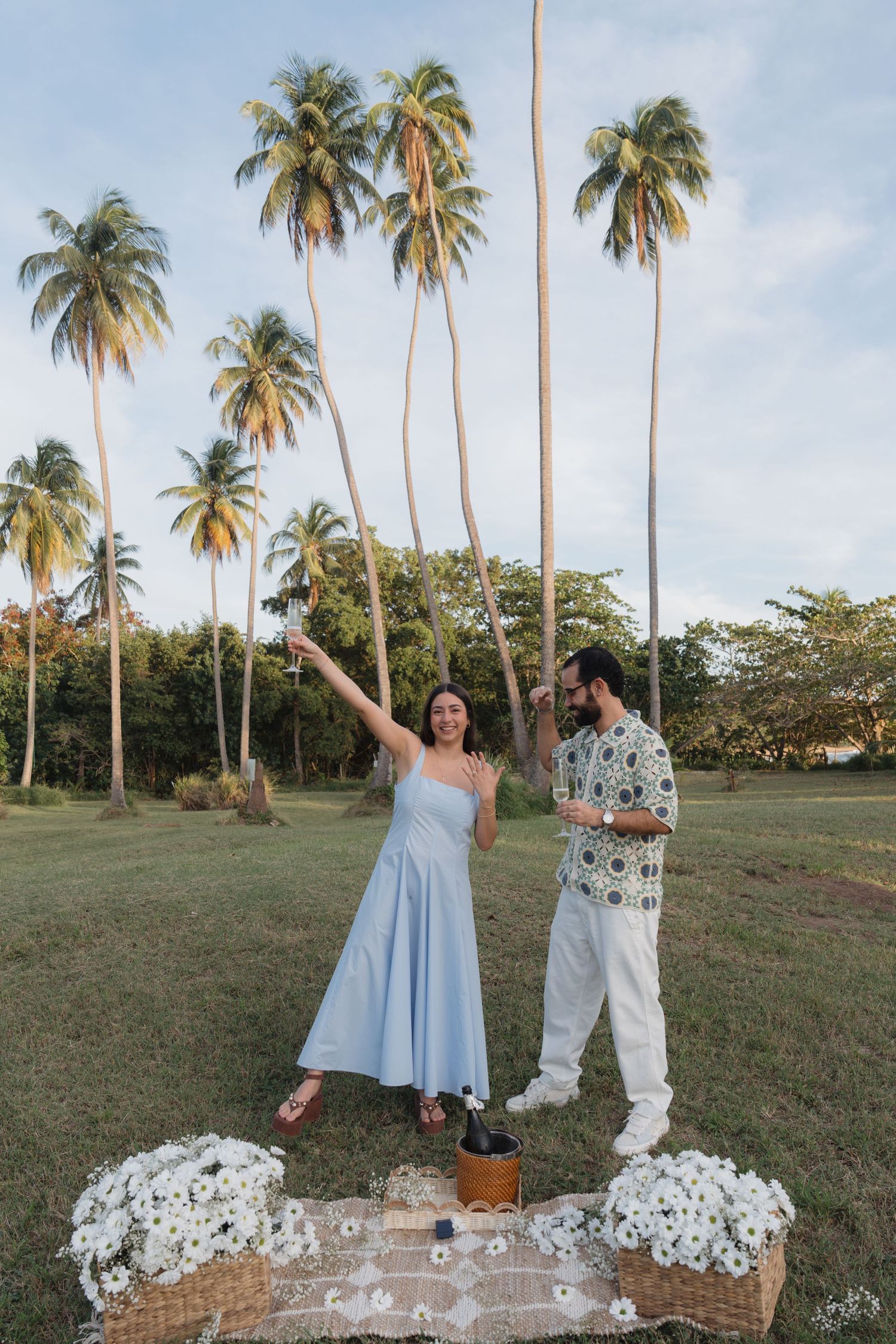 Couple celebrating outdoors under tall palm trees with white flowers and a picnic blanket on green grass.
