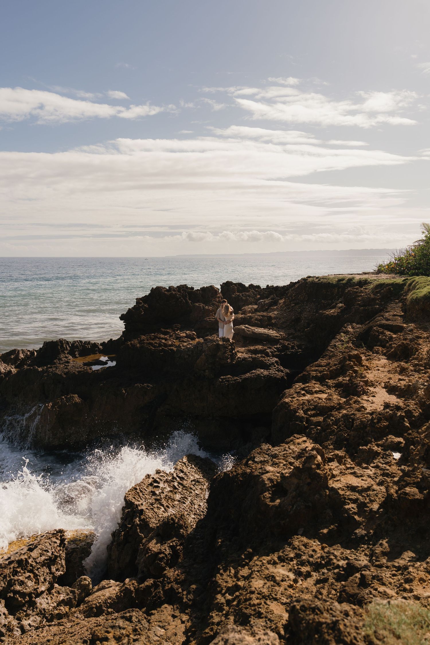 A person stands on rugged volcanic rocks by the ocean as waves crash below under a partly cloudy sky.