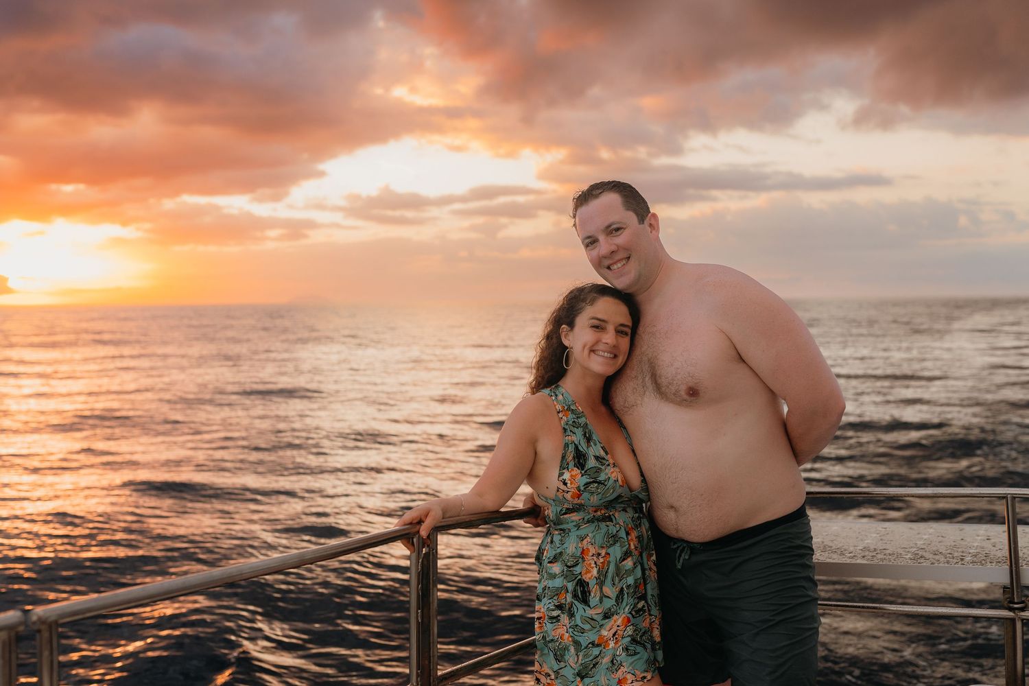Couple smiling together on a deck at sunset over the ocean, with vibrant orange and pink skies.