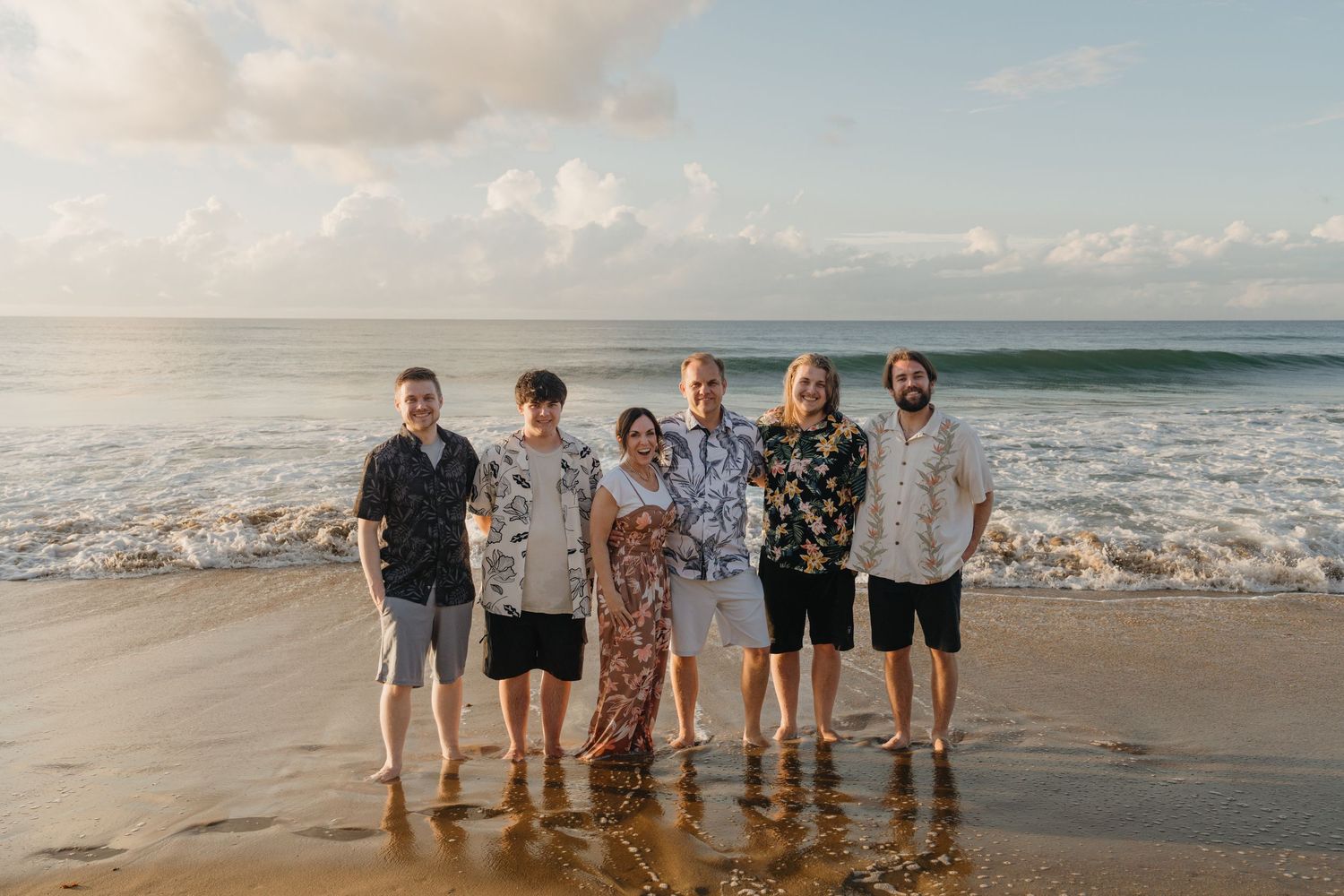 Six people smiling together on a sandy beach at sunset with ocean waves and cloudy sky in the background.