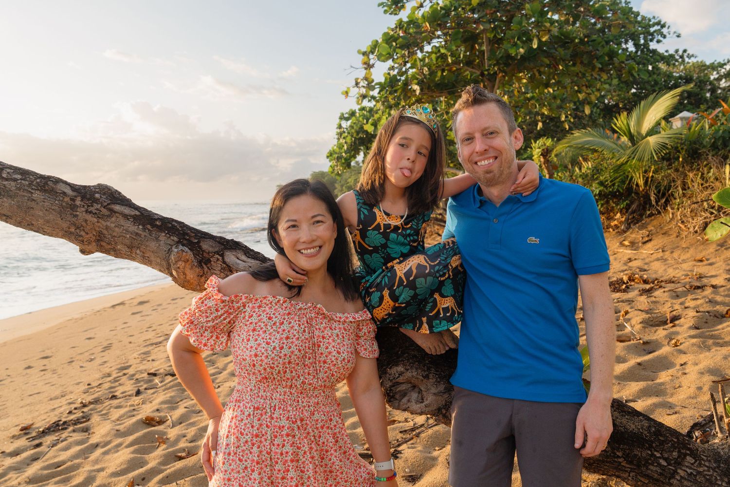 Family of three smiling on a tropical beach at sunset, posing near a fallen tree with lush greenery.