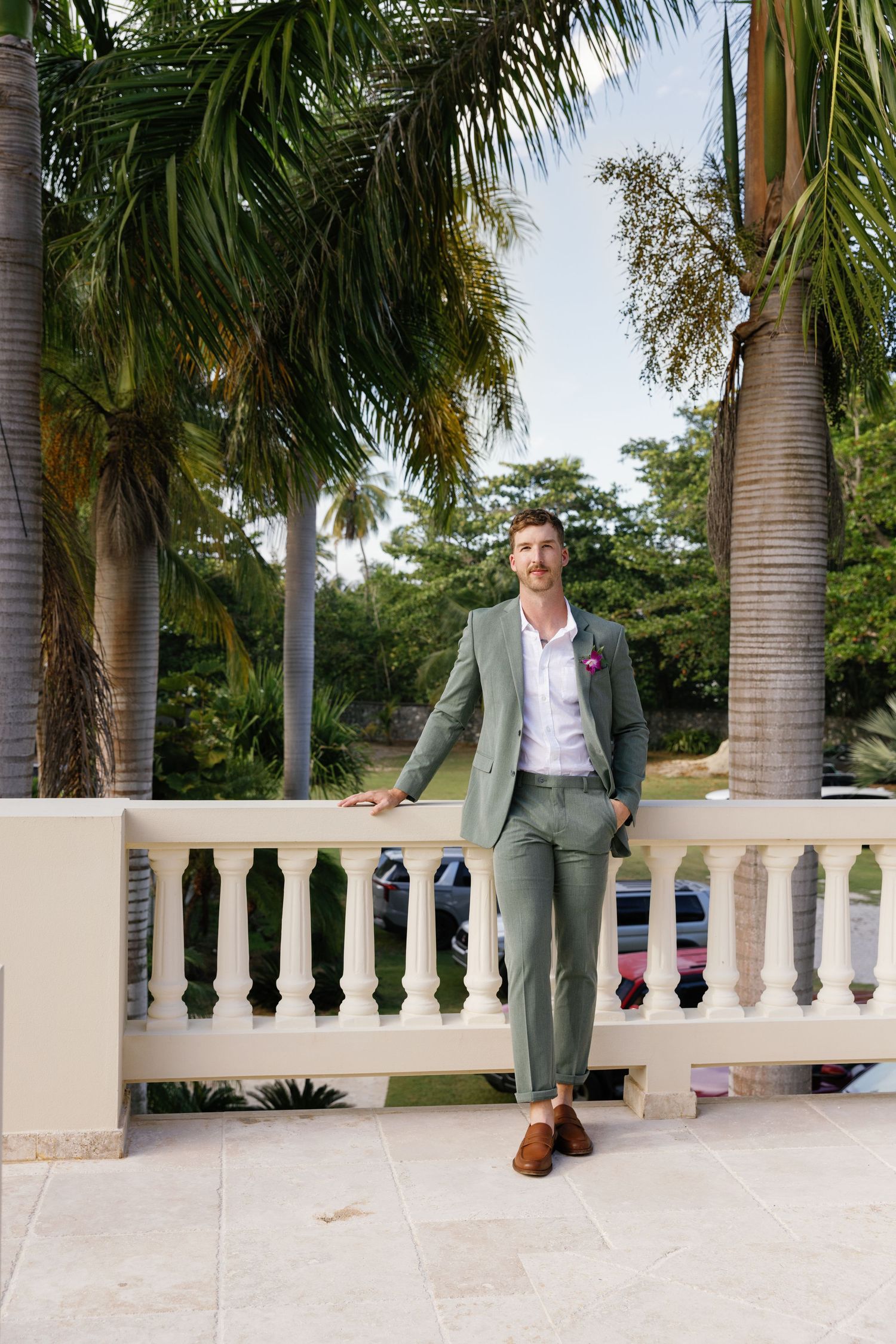 Groom in sage green suit leaning on white balustrade with tropical palm trees in background.