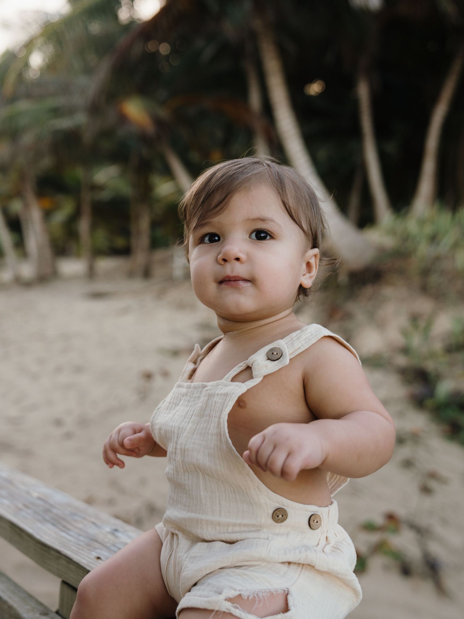 Adorable baby in cream linen romper sitting on wooden bench at sandy beach with palm trees.