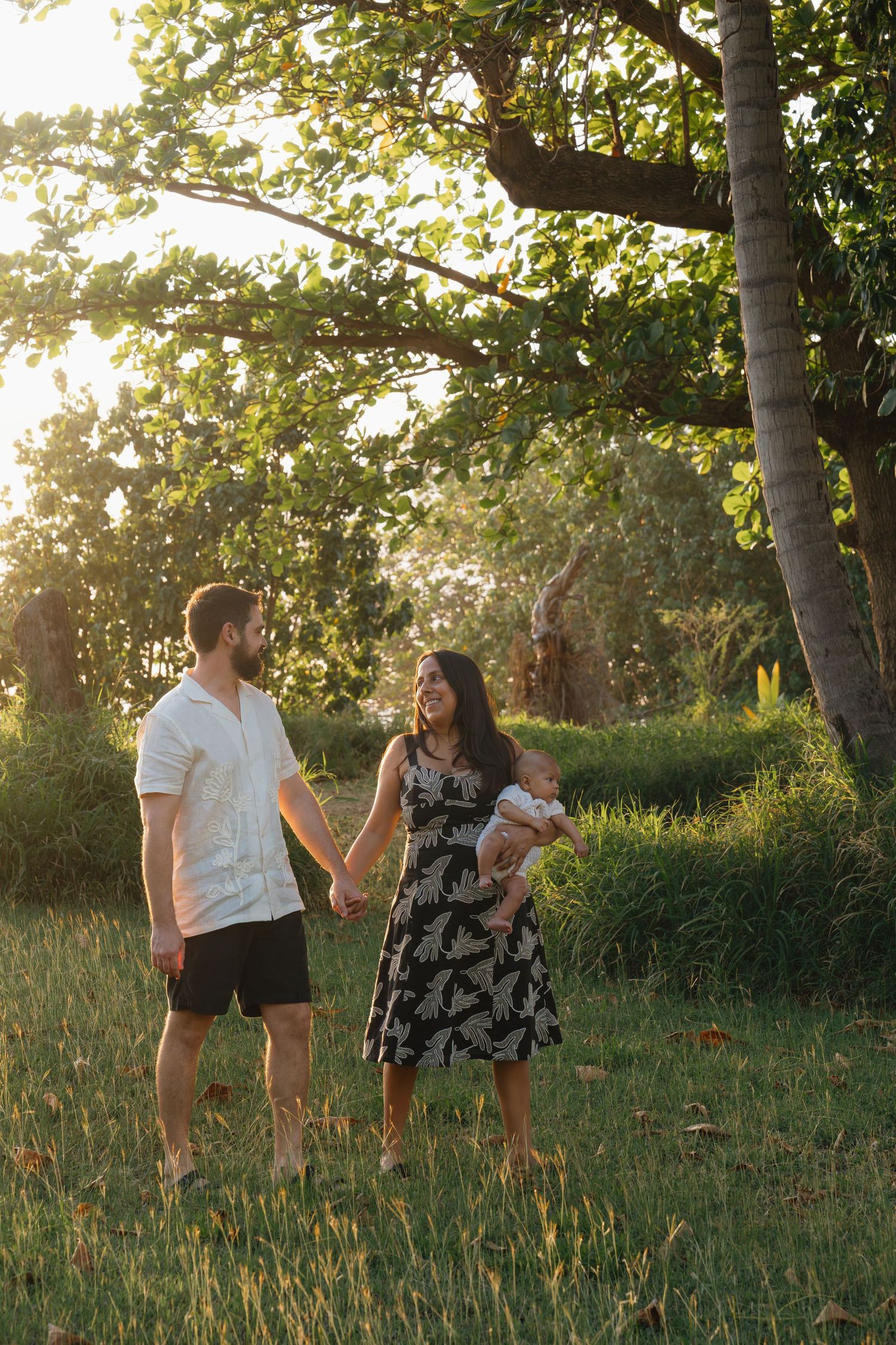 A couple holds hands in a sunlit park, woman in floral dress holding a baby, golden hour glow through lush trees.