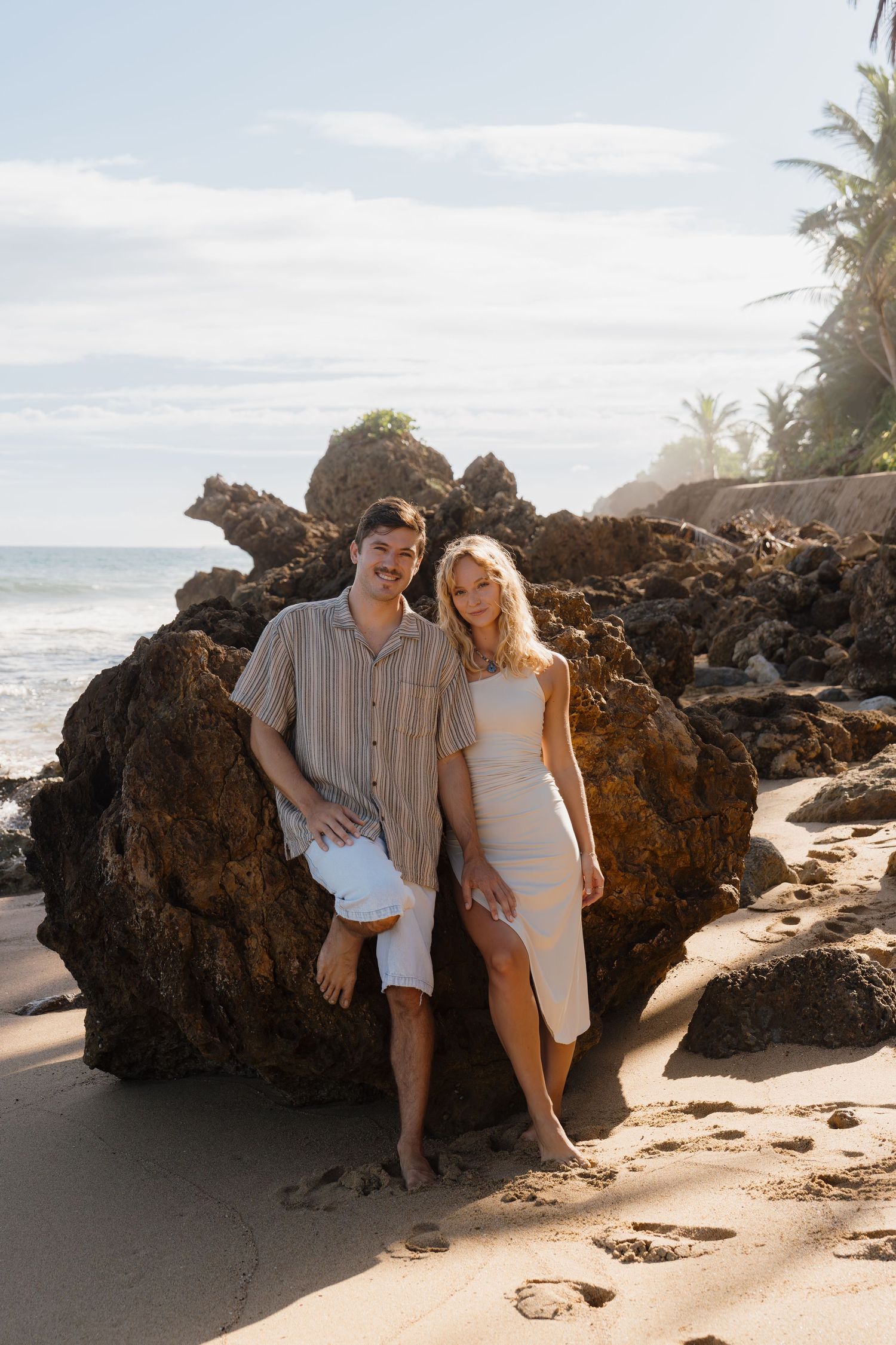 Couple posing together on a sandy tropical beach with rocky cliffs and palm trees in the background.