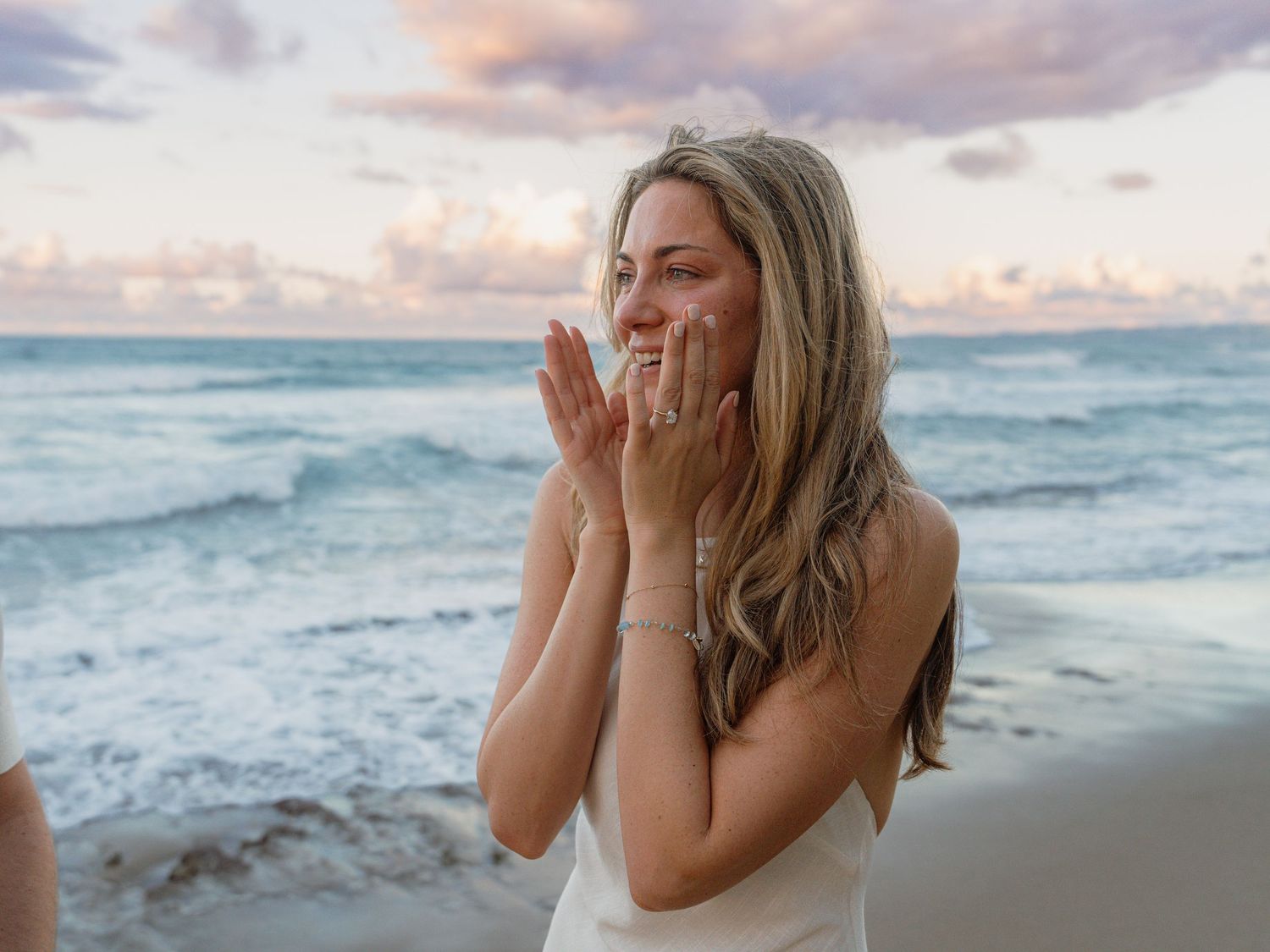 A joyful blonde woman in a white dress laughs on a beach at sunset, hands raised to her face.