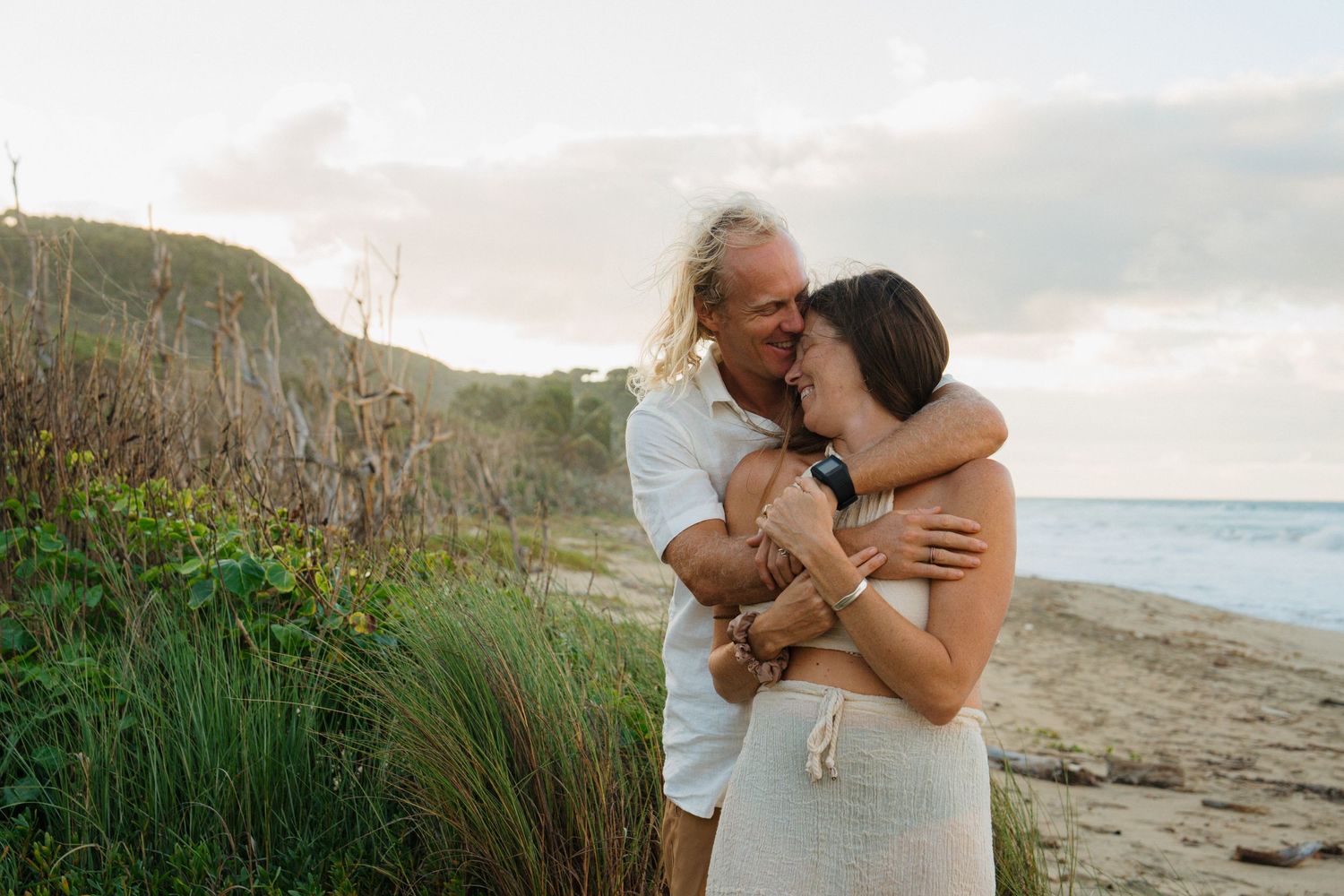 Couple embracing on a sandy beach at sunset, man kissing woman's head near coastal dunes.