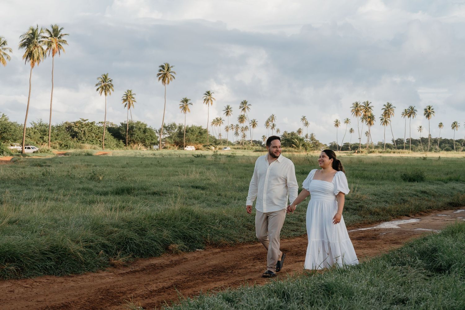 Couple in white outfits walking hand in hand on a dirt path surrounded by lush green fields and tall palm trees.