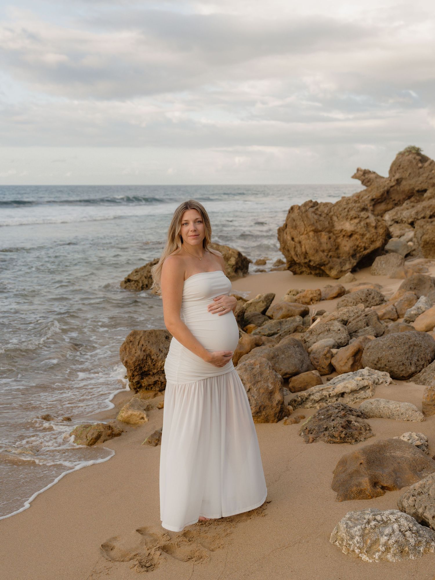 Pregnant woman in white strapless gown cradling baby bump on rocky beach with ocean waves at sunset.