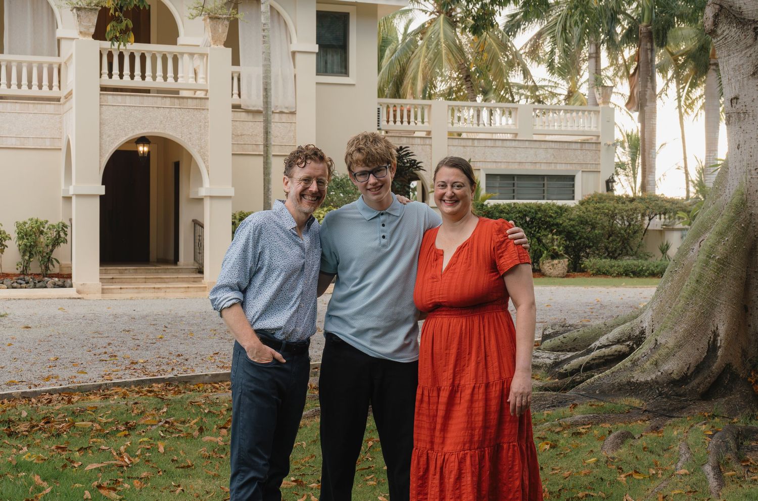 Three people smiling together outdoors in front of a tropical mansion with palm trees.