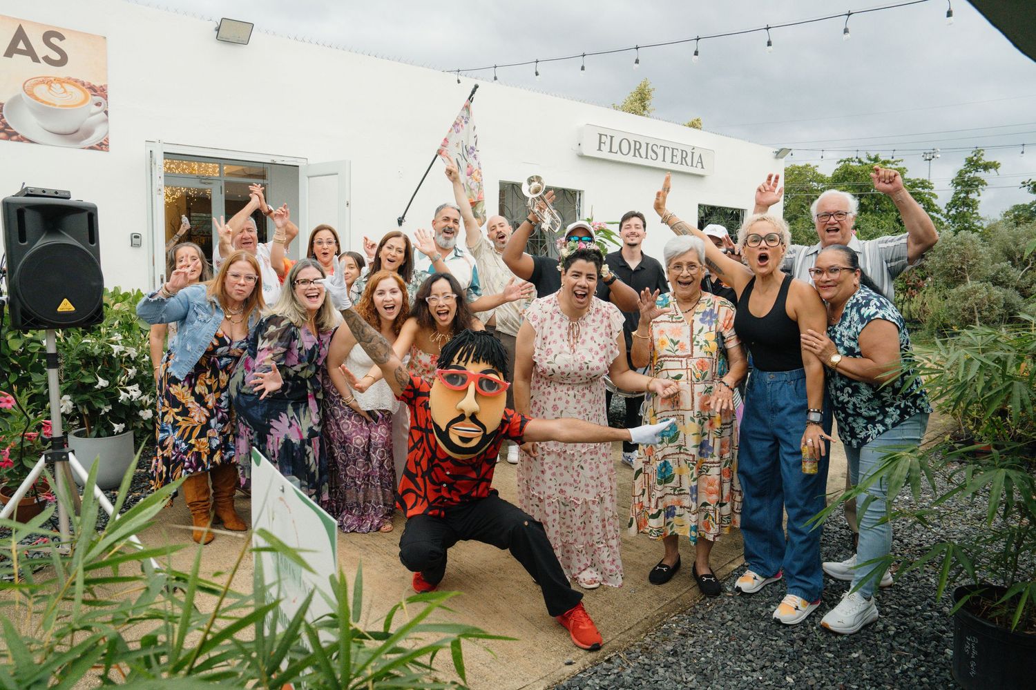 Joyful group celebration outside a florist shop with people cheering and a performer in a colorful mask posing energetically.