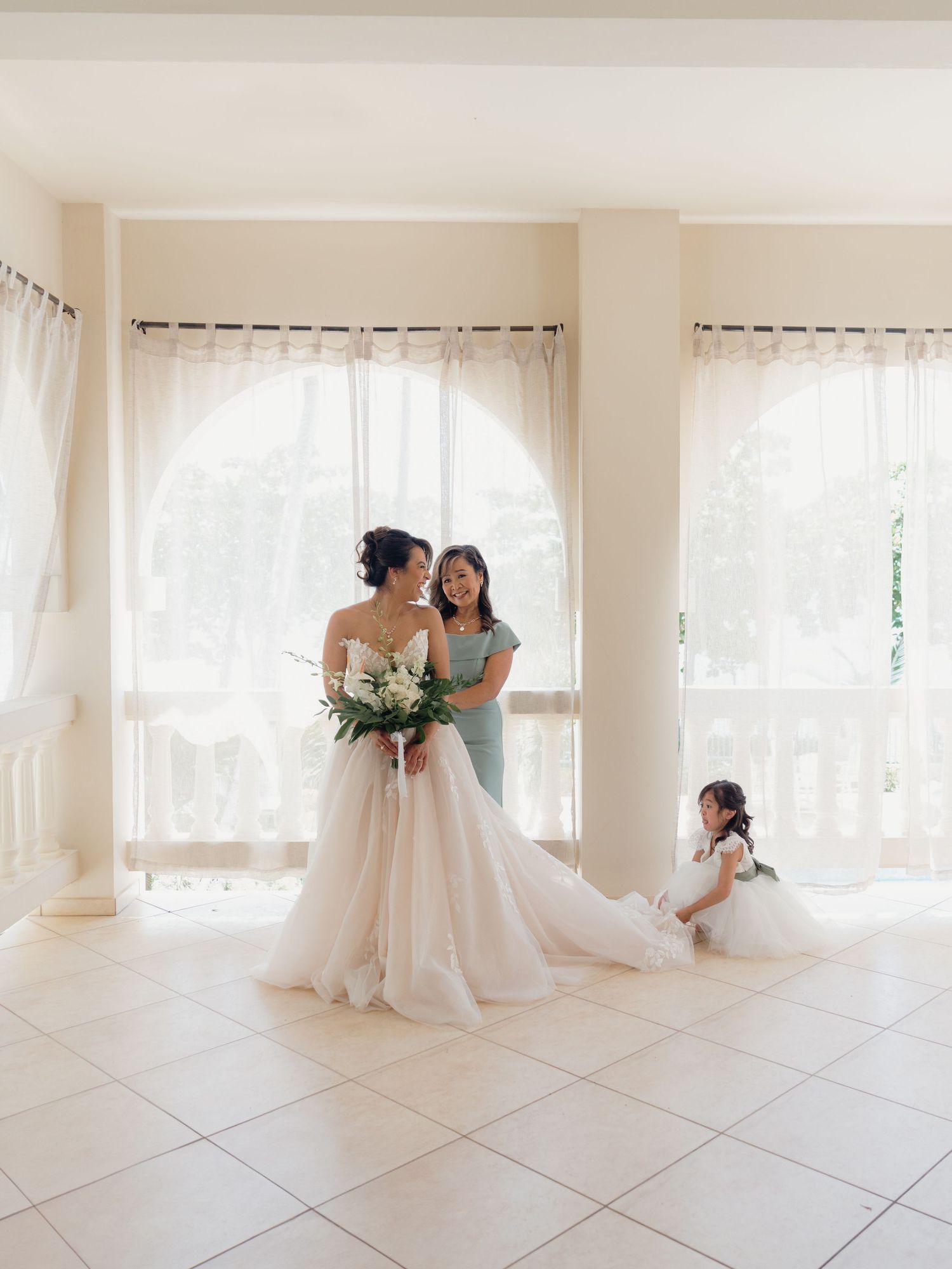 Bride in a blush tulle gown holds bouquet beside bridesmaid in sage green, with flower girl nearby in bright room.