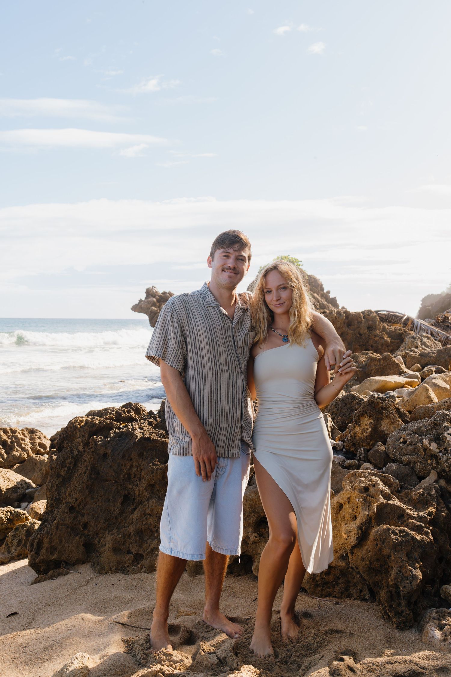 A smiling couple poses together on a rocky beach with ocean waves in the background on a sunny day.