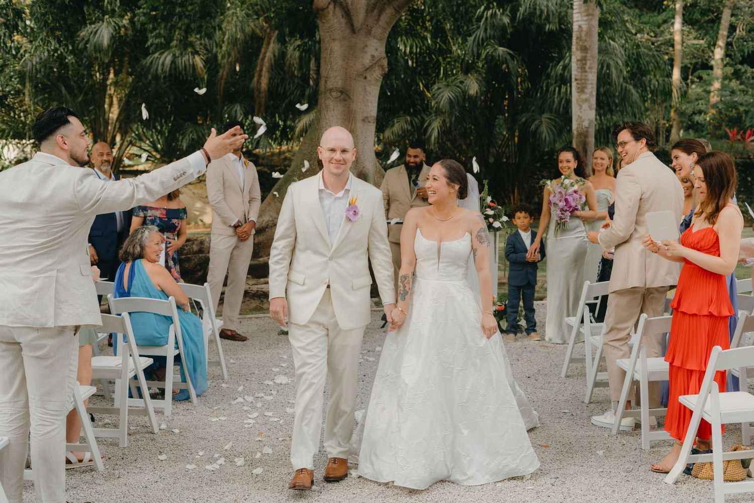 Newlywed couple walks down the aisle as guests toss flower petals at an outdoor tropical wedding ceremony.