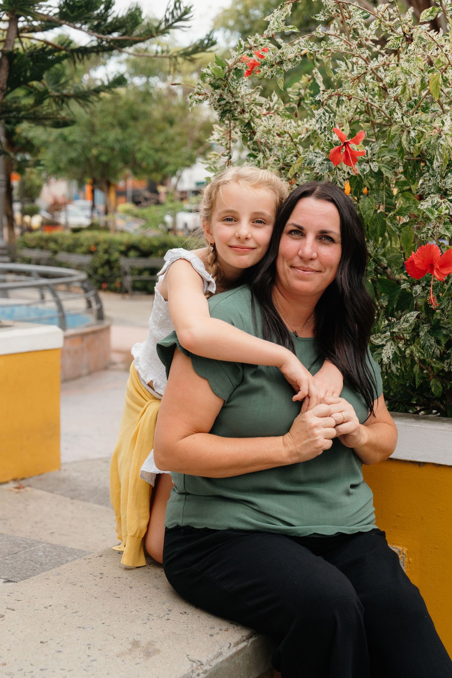 A woman and young girl share a warm hug outdoors near colorful flowers and greenery in a park setting.